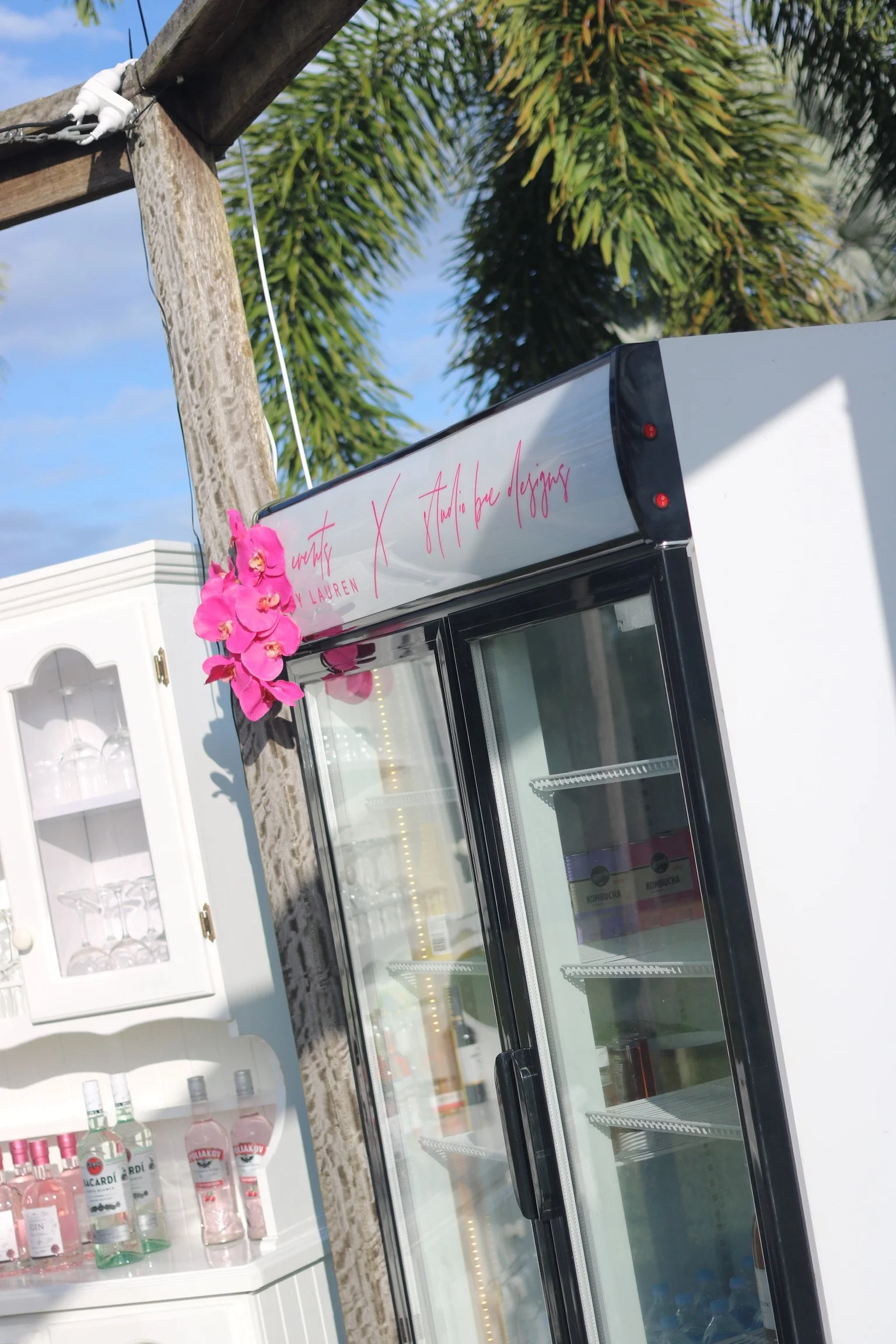 An outdoor bar setup featuring a white refrigerator, a white cabinet with glassware, and several bottles of alcohol, with a palm tree and a partly cloudy blue sky in the background.