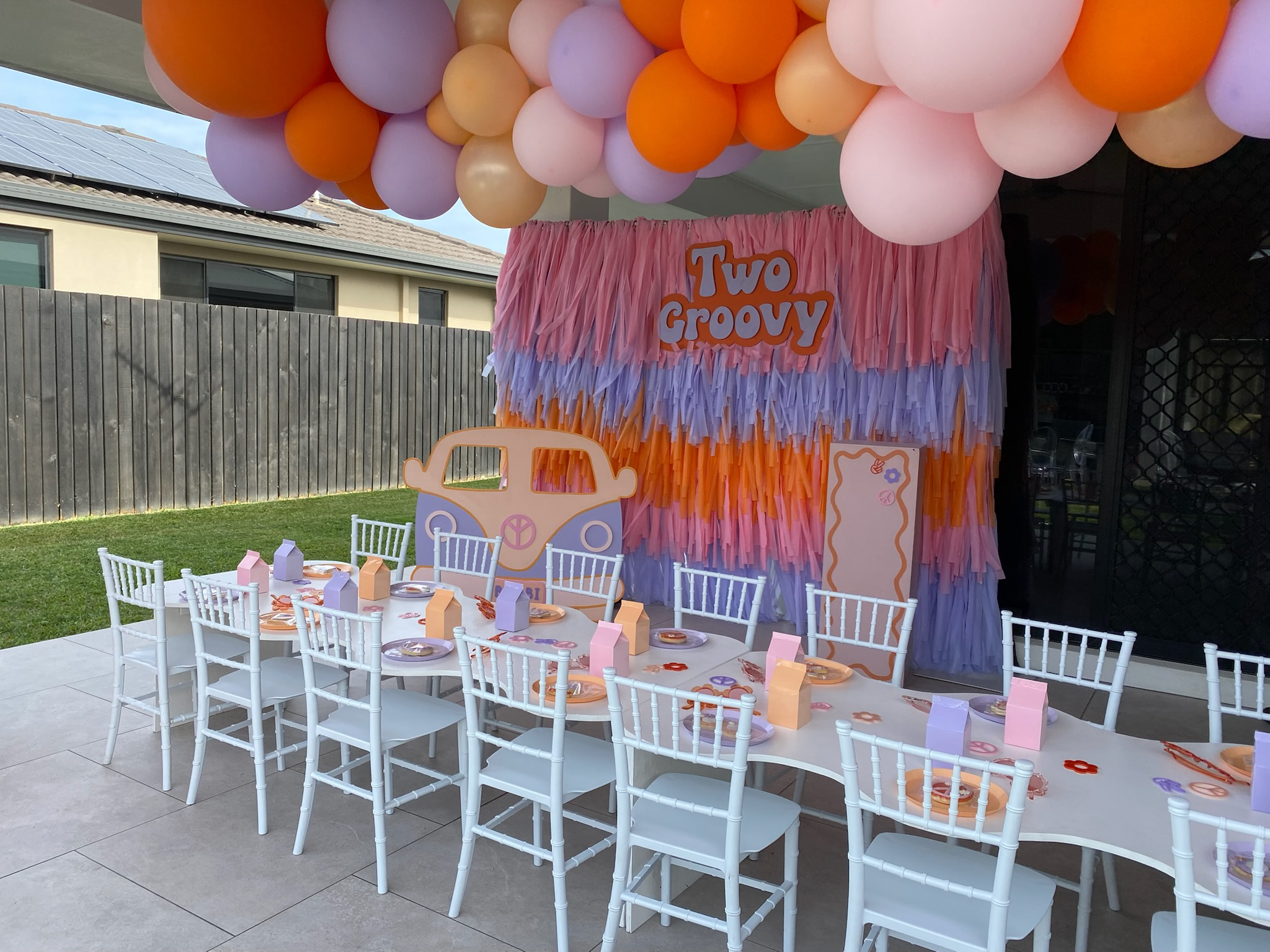 Decorated outdoor party area with a long white table surrounded by white chairs, set with plates and pink and purple party favor boxes. Behind the table, there is a pink, purple, and orange fringed backdrop with a large sign that reads 'Two Groovy' a