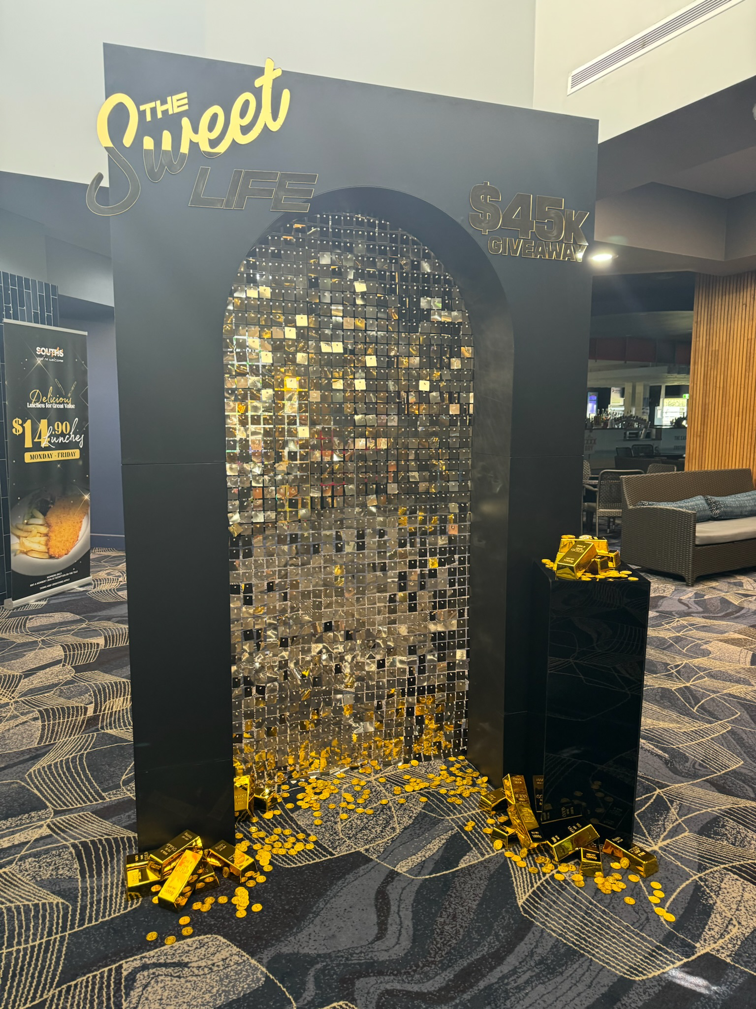 Black display stand with golden coins and gold-colored bars for a promotion, with decorative mirror tiles and signage advertising a $45,000 giveaway.