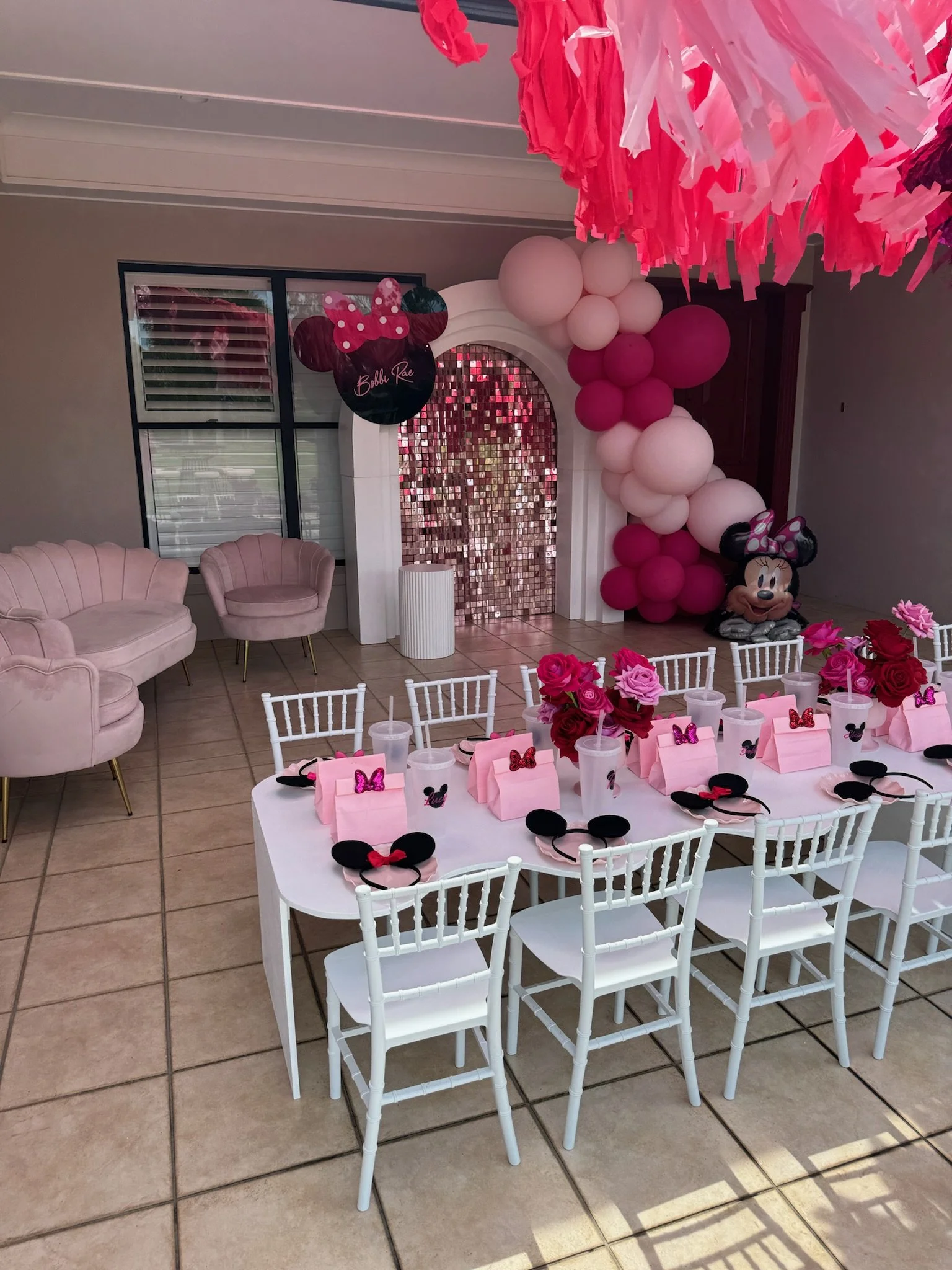 A decorated indoor party setup for a children's birthday with a pink and Minnie Mouse theme. A table with pink gift bags, Minnie Mouse headbands, and pink roses in vases. Pink and white balloon arrangements and Minnie Mouse balloons on the walls, wit