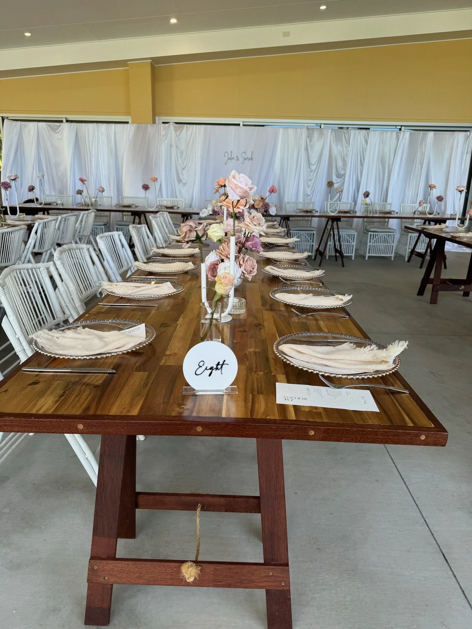 Elegant wedding reception table with pink and white floral centerpieces, clear glass plates, beige napkins, and white chairs. White draped backdrop with floral arrangements and a sign that says "Jale & Sarah".