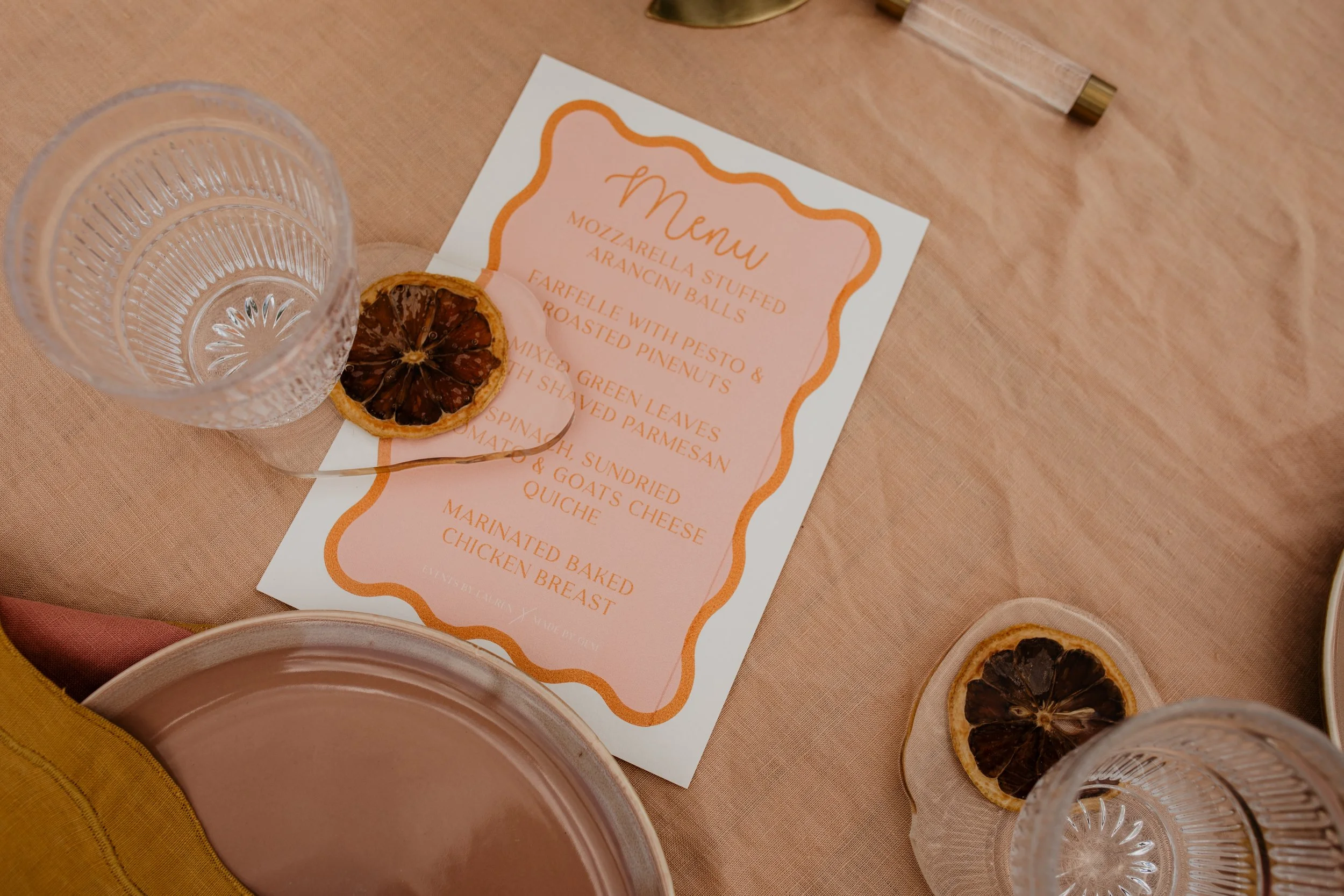 A table setting with two clear glass cups with dried citrus slices on top, a printed menu with orange and pink accents, a golden pen, and a partially visible plate with a yellow cloth underneath.