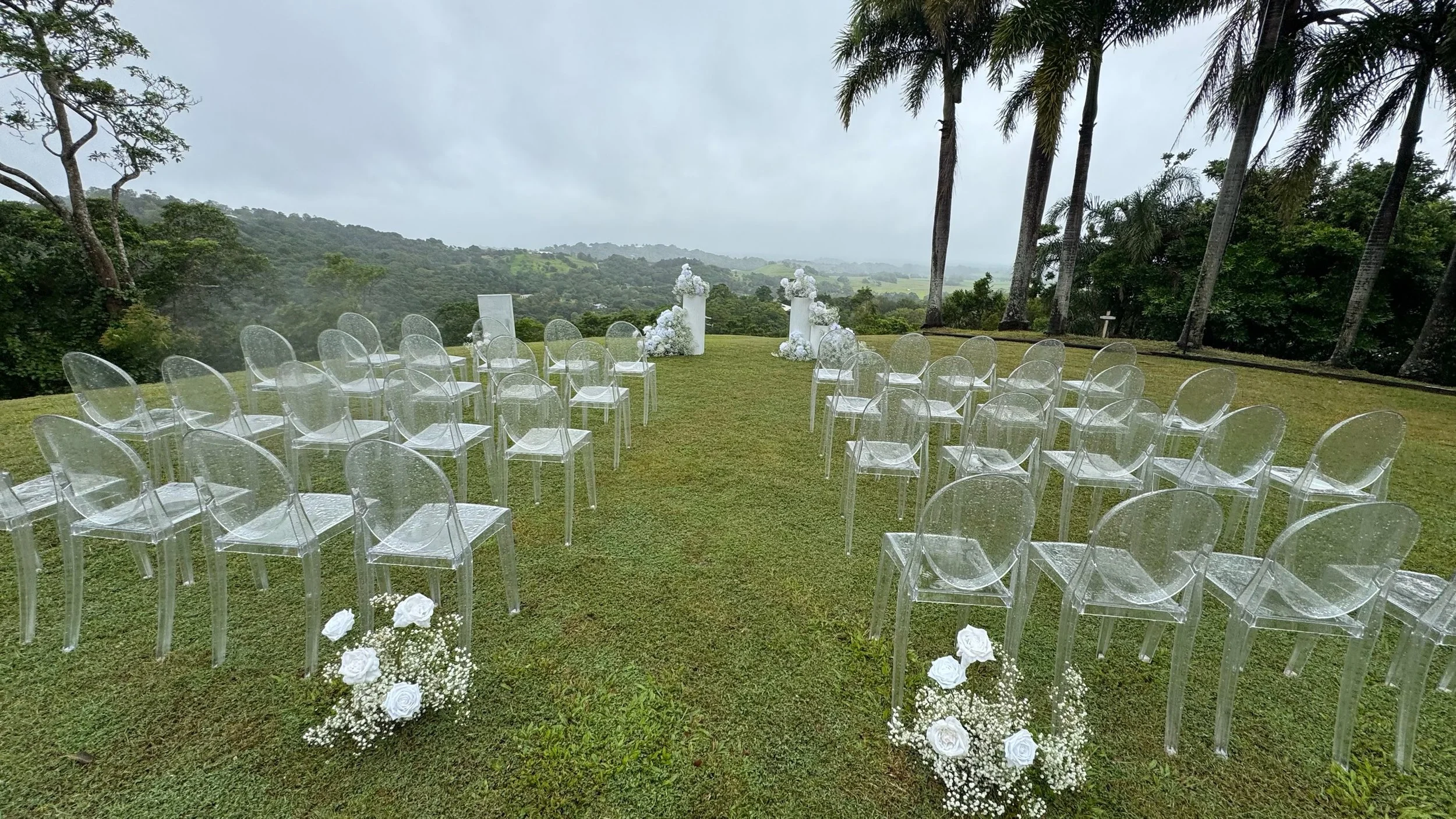 Outdoor wedding ceremony setup with transparent chairs arranged on grass leading to a floral arch, surrounded by trees and hills under cloudy sky.