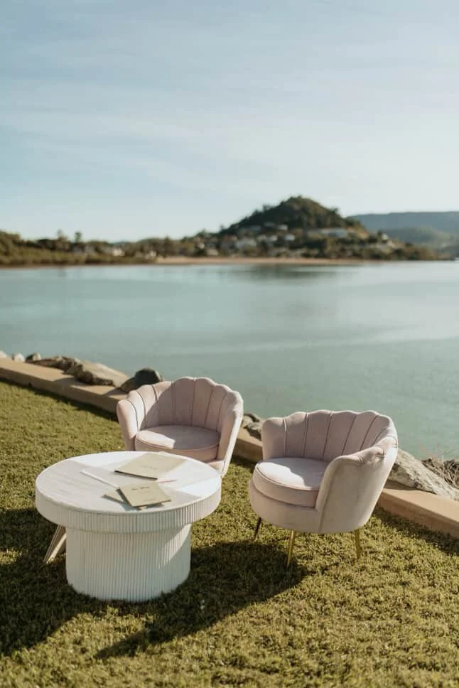 Two pink velvet armchairs and a white round table with books, set near a body of water with hilly landscape in the background.