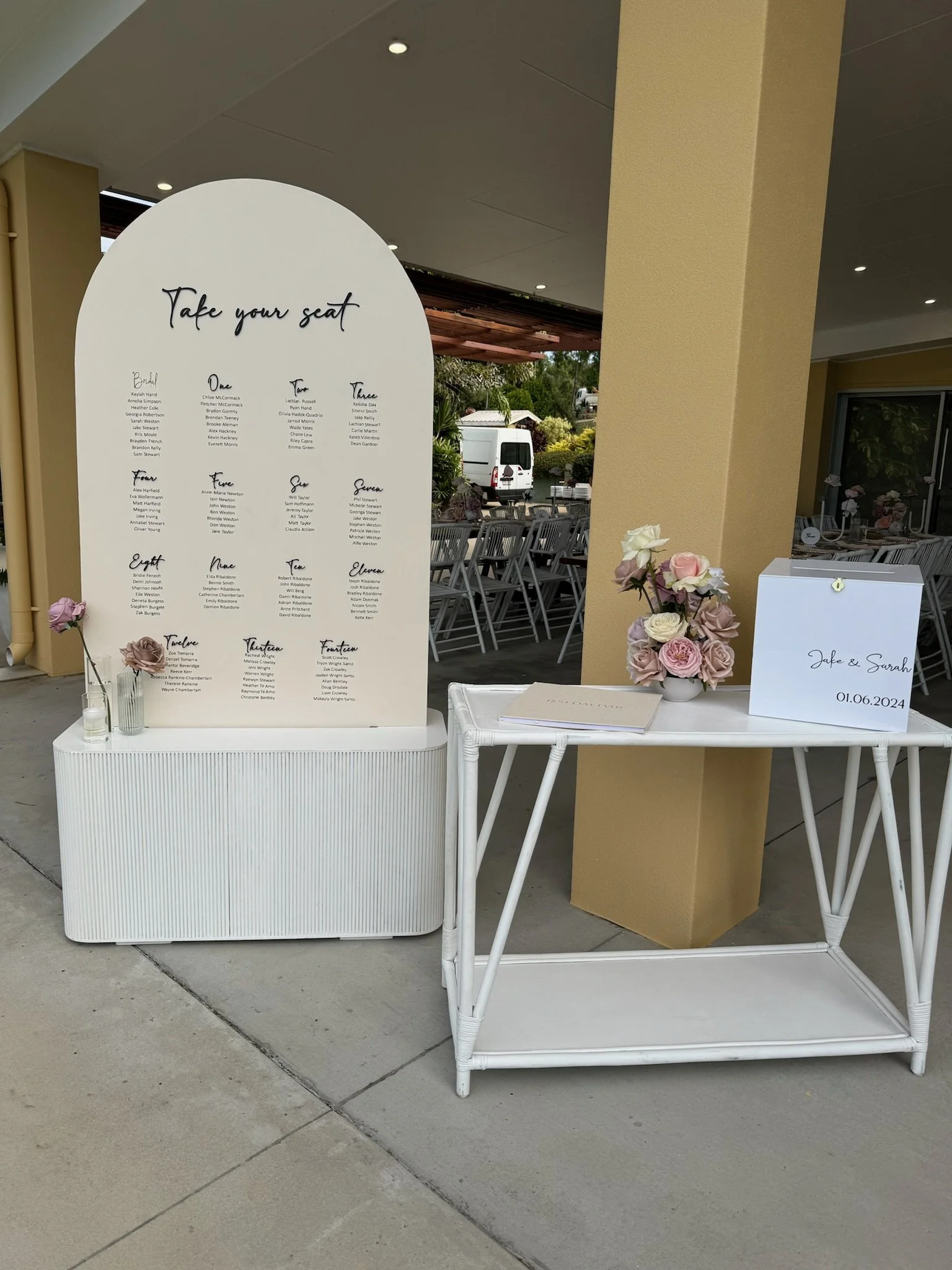 Wedding seating display with a large white board listing table assignments, decorated with pink and white flowers, and a small white box with the names Jake and Sarah and the date June 1, 2024.