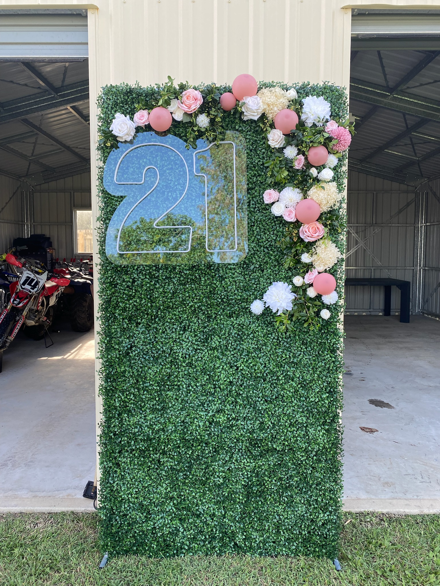 Green hedge wall decorated with pink and white flowers, pink balloons, and a large number 21 sign.
