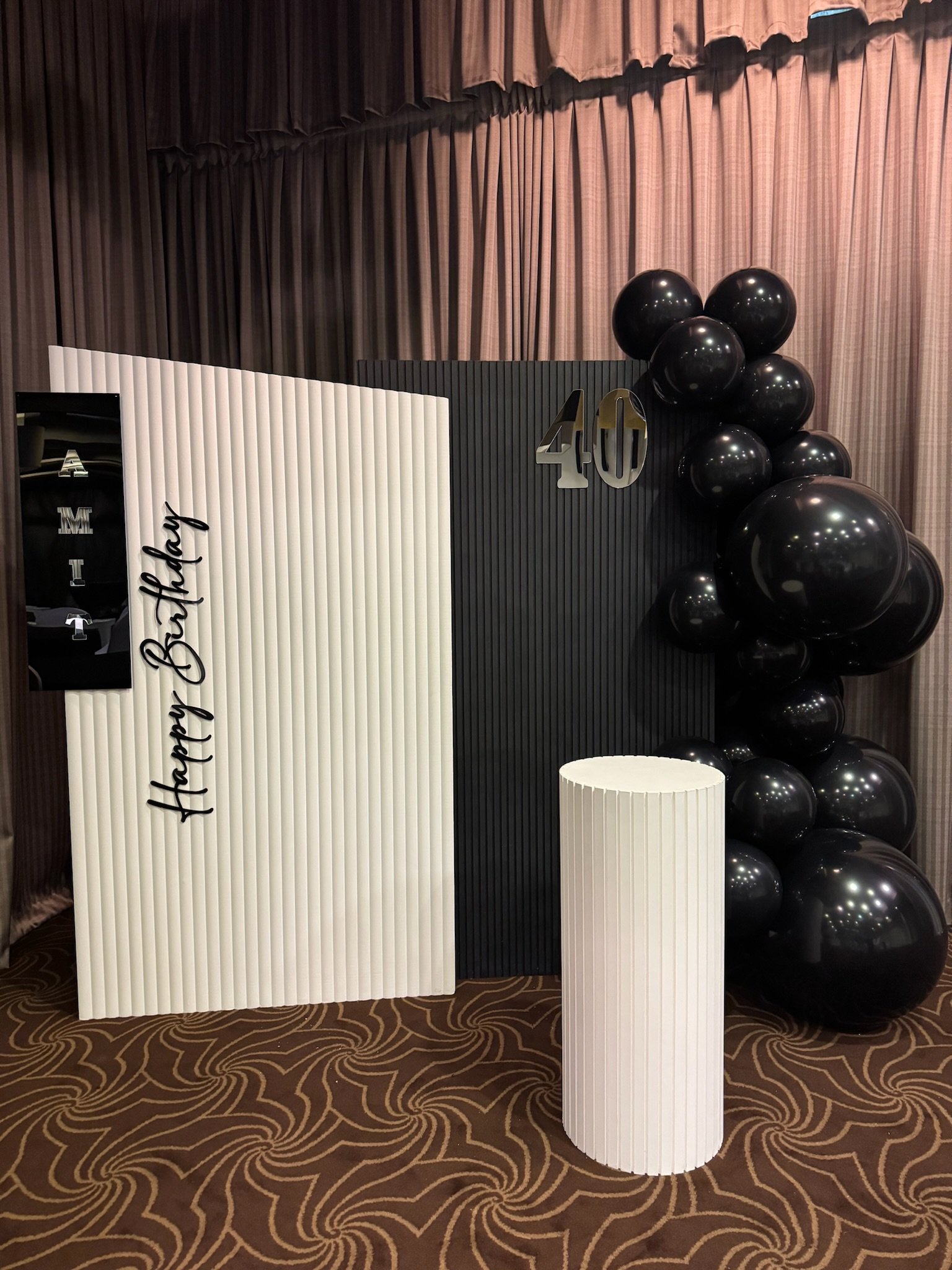 Decorative backdrop with black and white elements, featuring balloons, a sign reading 'Happy Birthday,' and the number 40, set on a patterned carpeted floor.