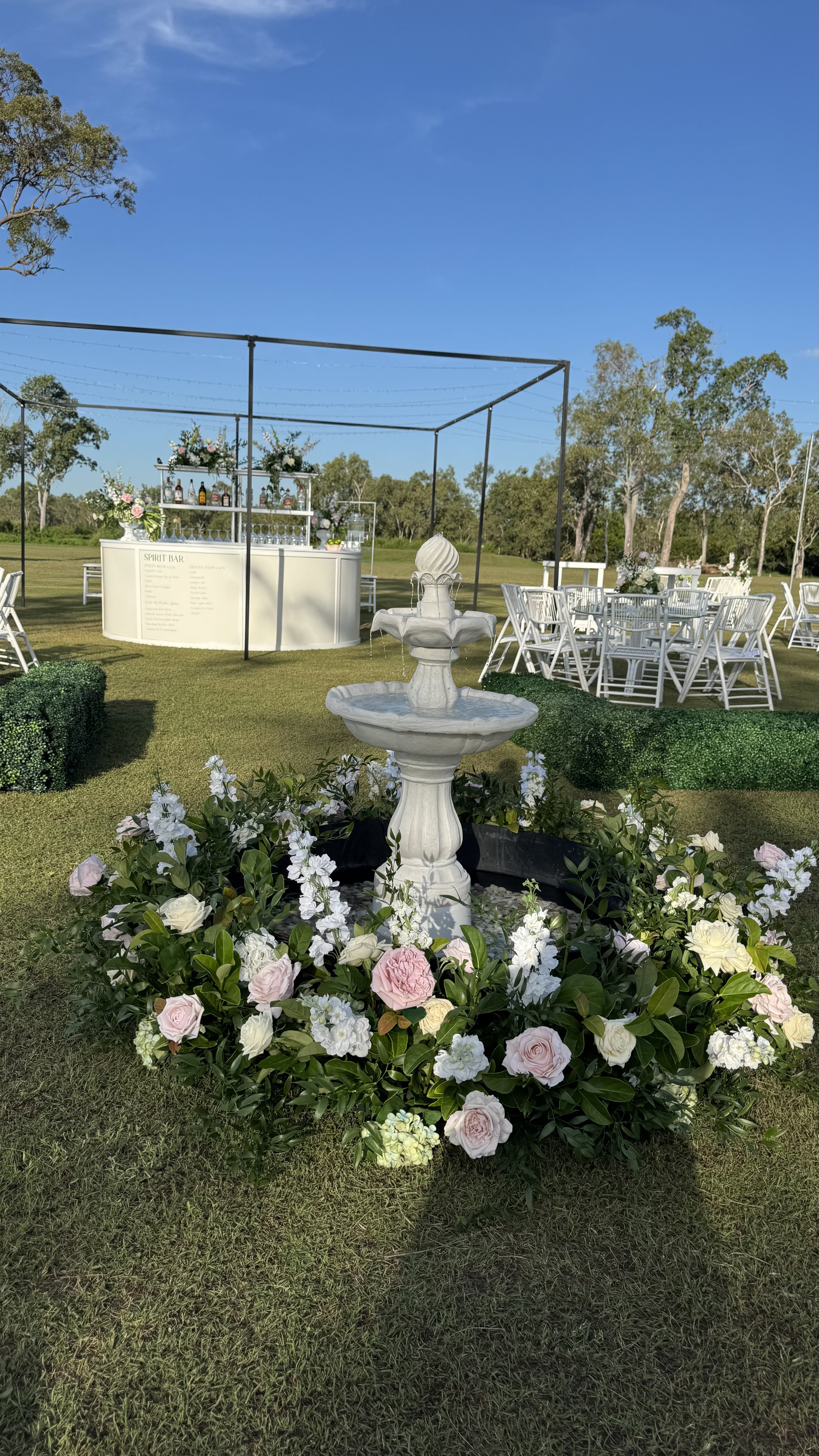 Outdoor event setup with a white fountain surrounded by pink and white flowers, white chairs and tables, and a bar in the background under a clear blue sky.
