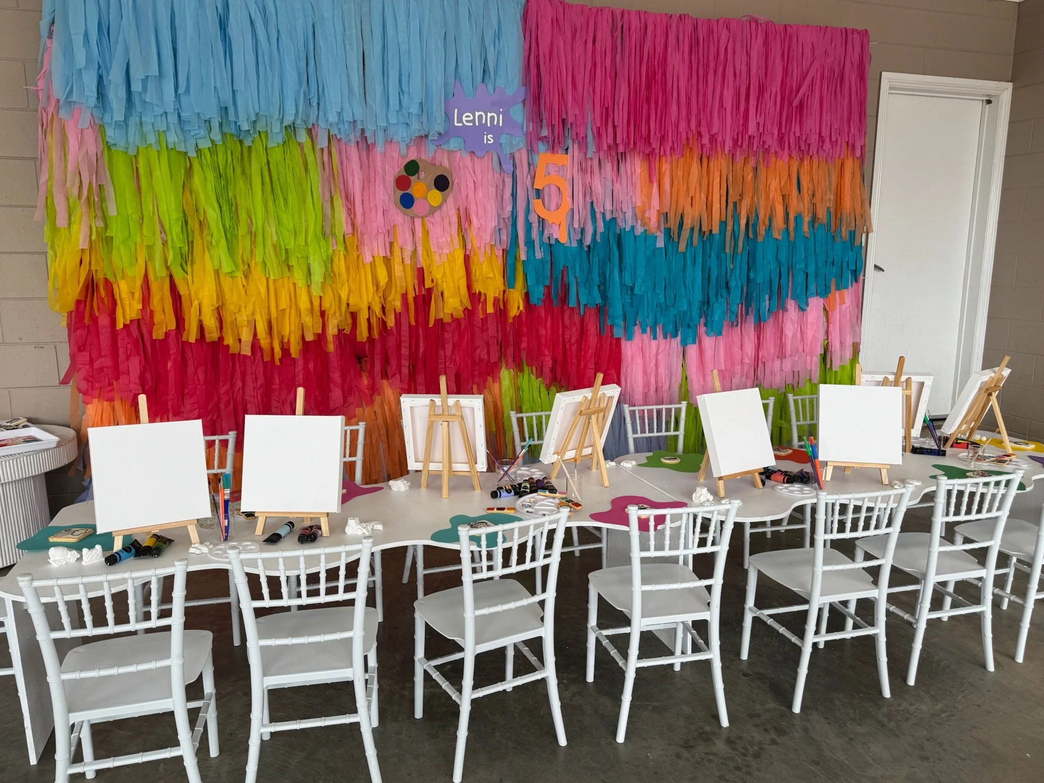 Colorful decorated party backdrop with white tables and chairs set up in front, ready for a children's art activity