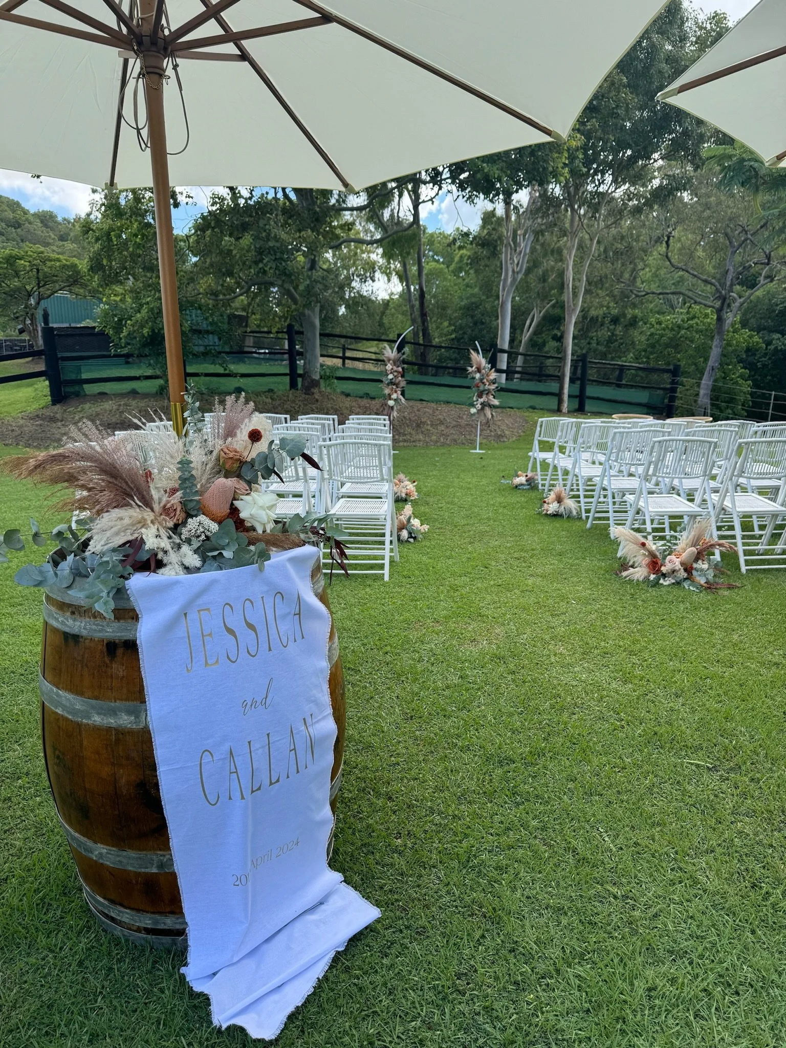 Outdoor wedding ceremony setup with white chairs, floral arrangements on the ground, a table with a cloth that reads "Jessica and Callan, April 2024," and two large umbrellas providing shade in a green, grassy area surrounded by trees.