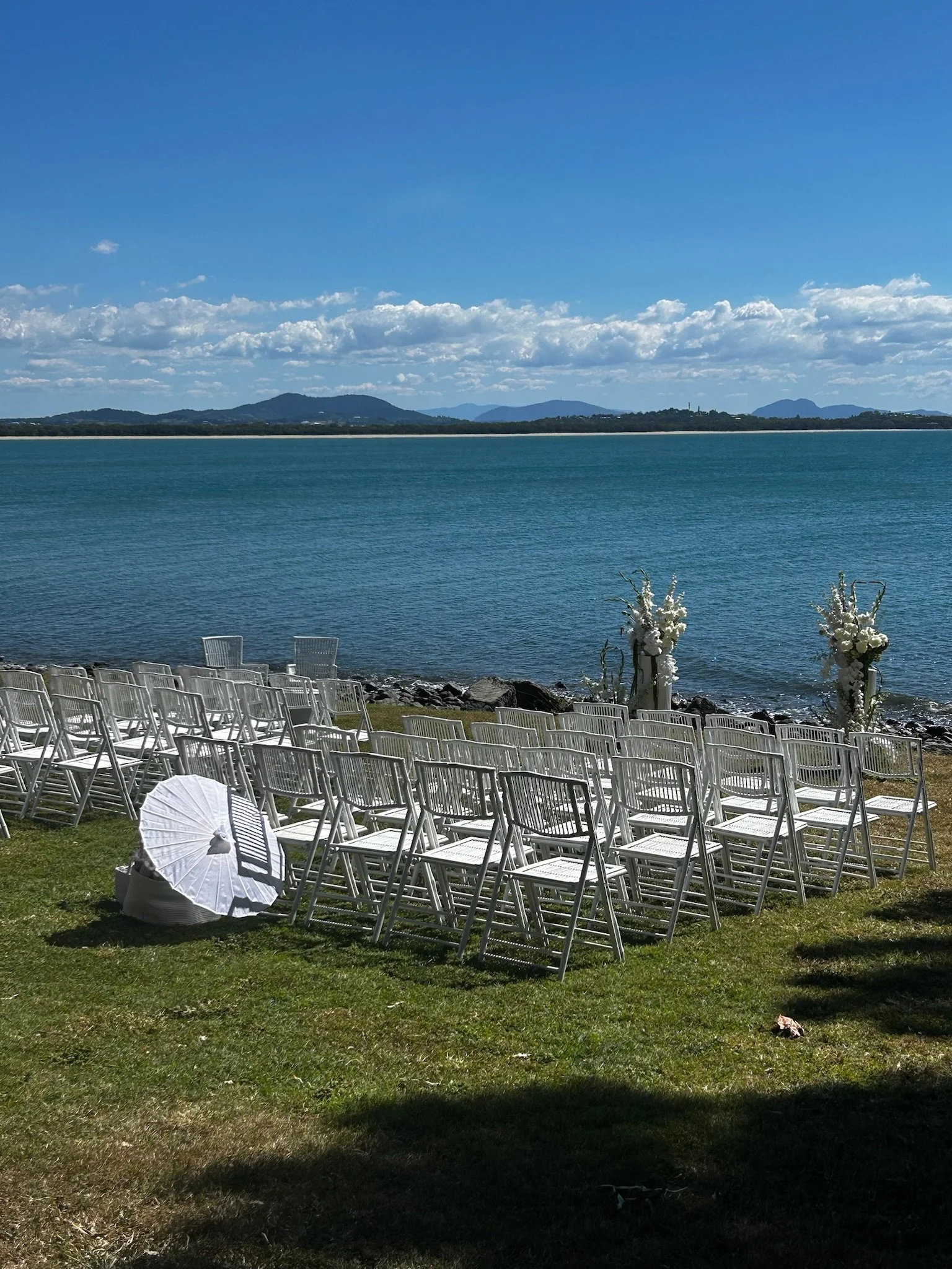 Outdoor wedding setup on the grass by a lake with white folding chairs, floral arrangements, and a white parasol, with mountains and a blue sky in the background.