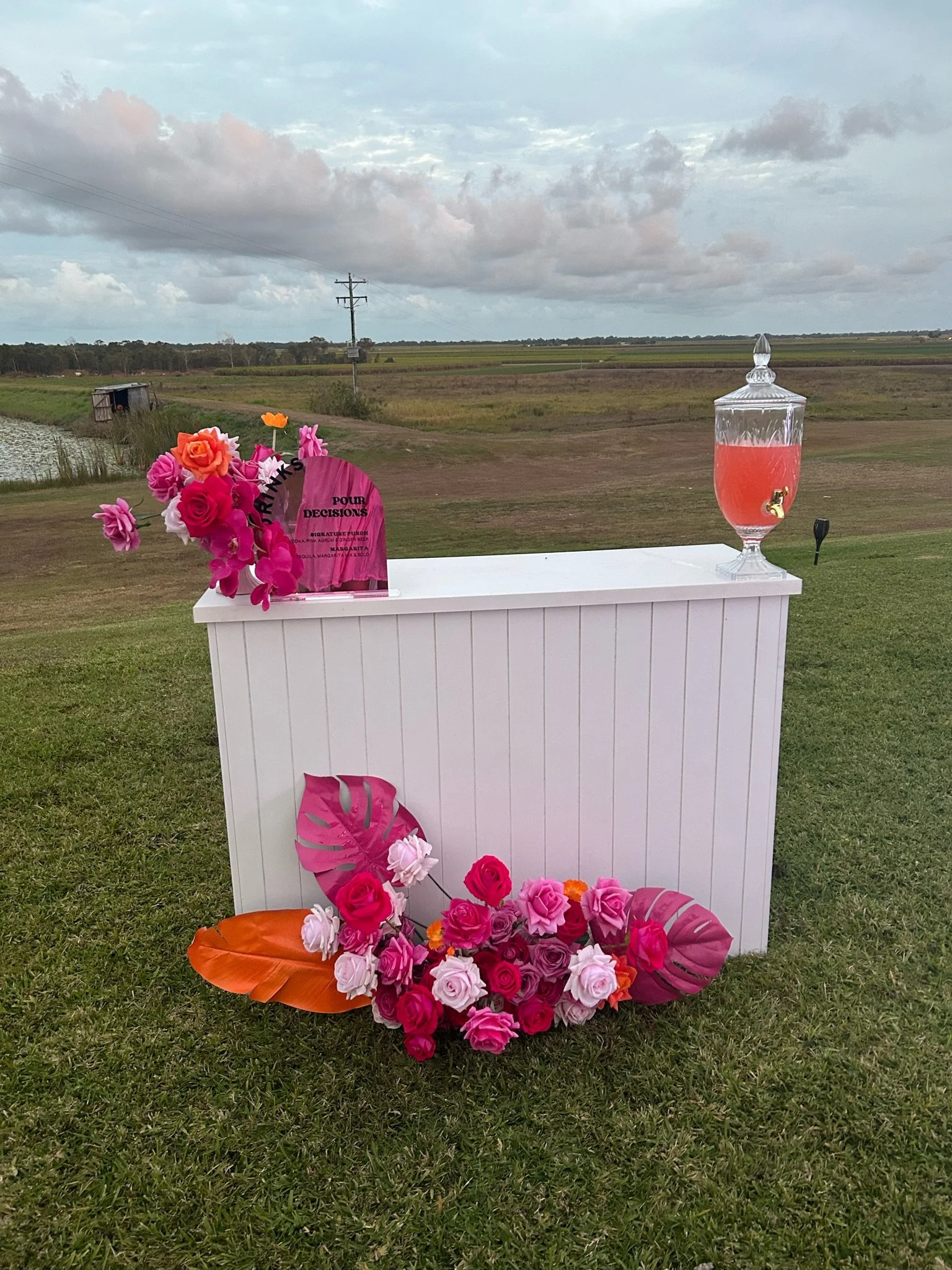 A white outdoor bar with pink and orange flowers, a pink drink in a glass container with a tap, and a sign that says 'Pour Decisions' in a scenic grassy area with cloudy sky and water in the background.