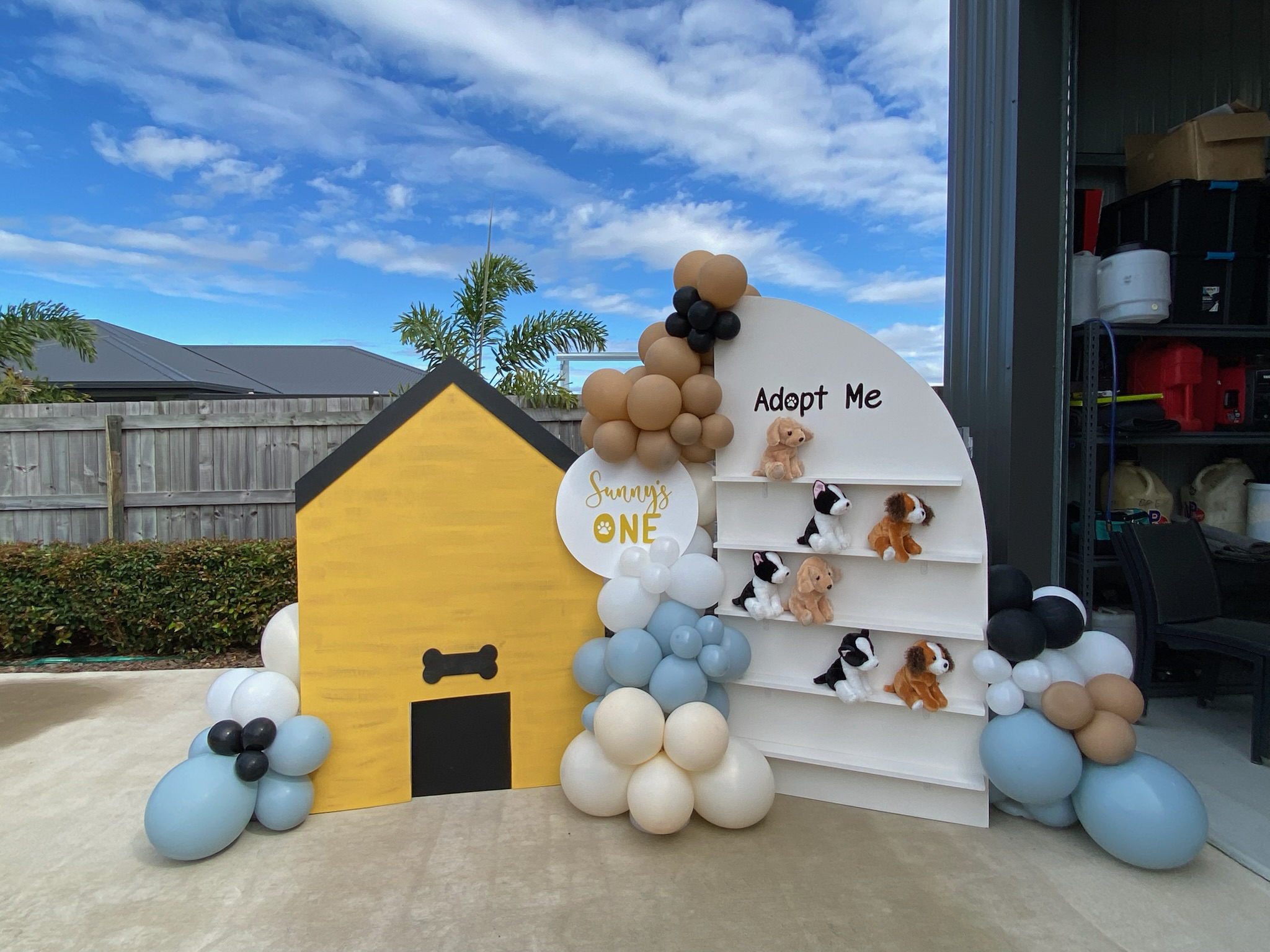 Dog themed birthday decoration featuring a yellow doghouse, balloons in neutral tones, and a white shelf with plush puppy toys and the words "Adopt Me".