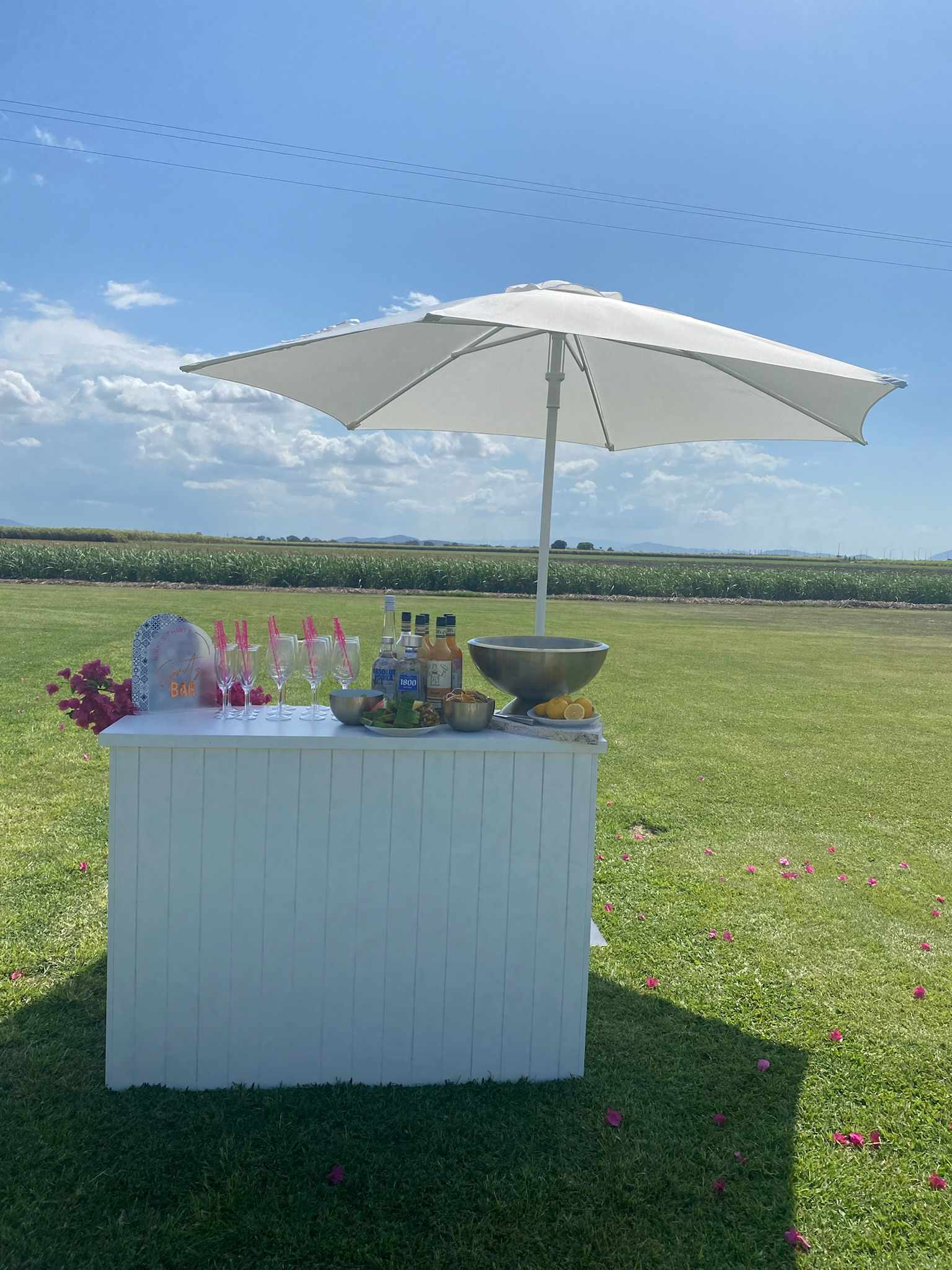 A white outdoor bar with a large umbrella, set on a grassy field with pink flower petals scattered around. The bar has glasses, condiments, a bowl of lemons, and a decorative sign that says 'BAR'.