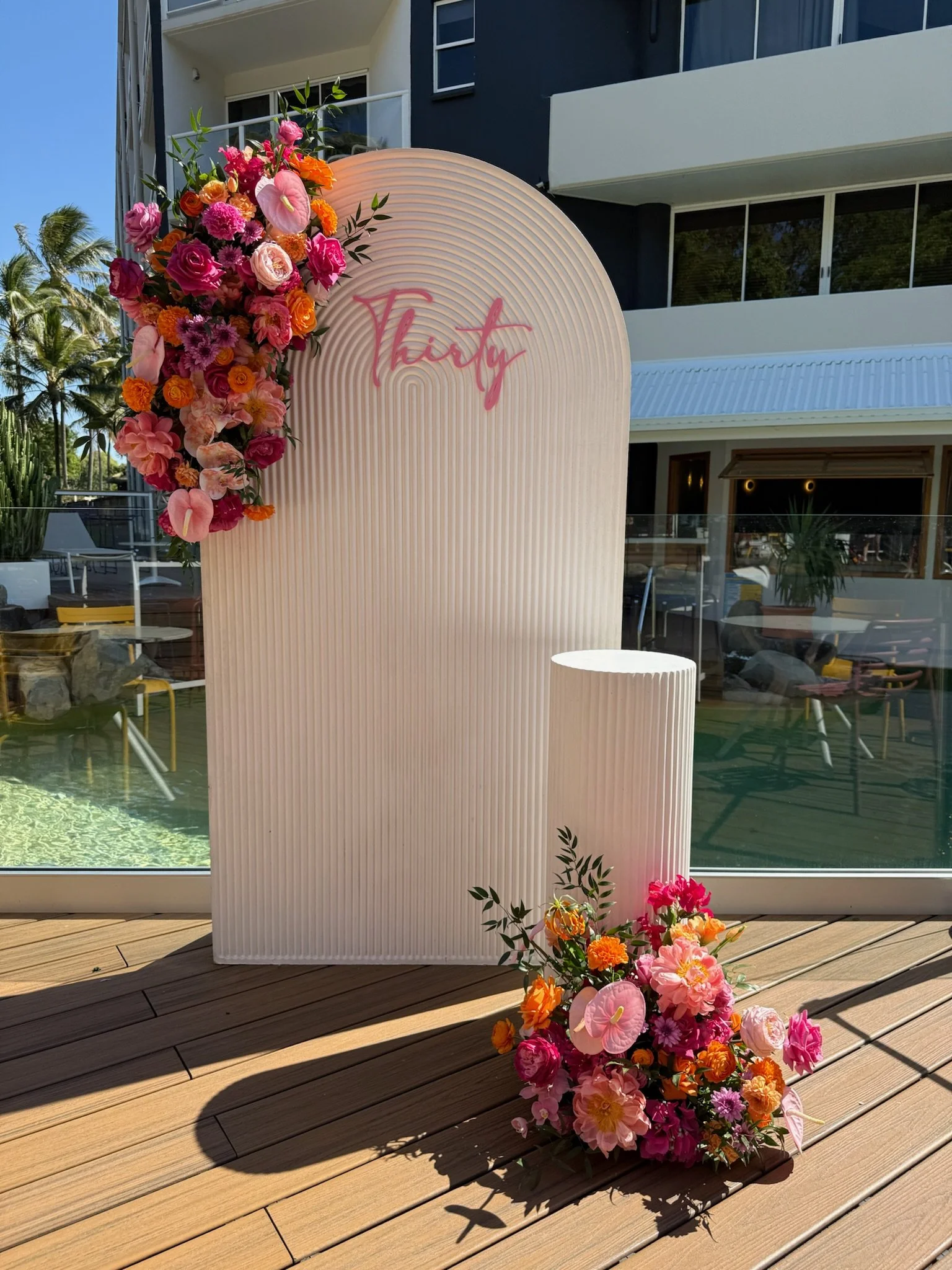 A decorative setup with a white textured panel, pink "Thirty" sign, and colorful flower arrangements in pink, orange, and purple, outdoors on a wooden deck near a glass building.