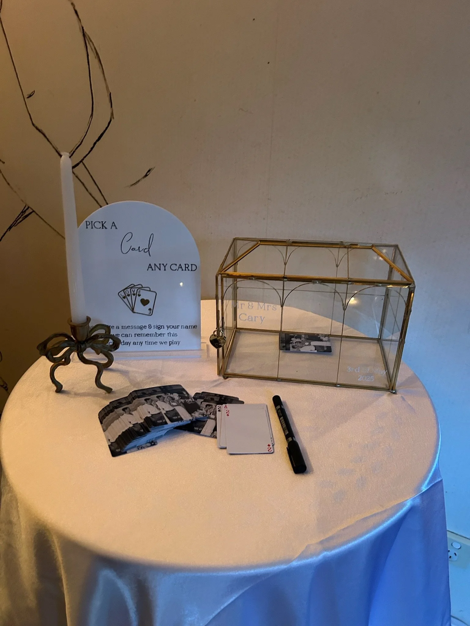 A table with a candle, a sign instructing guests to pick a card, several playing cards fanned out, and a glass box for cards. There is also a marker pen and a small card inside the box. The sign states: 'Pick a card, any card. Leave a message & sign 