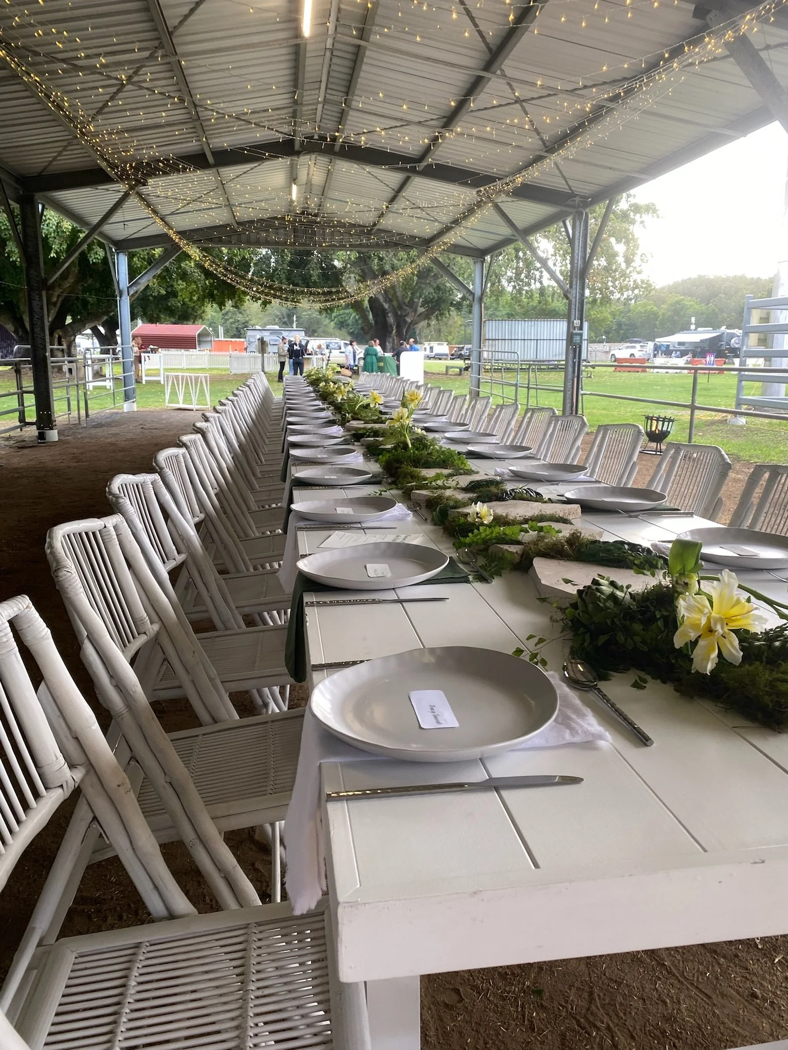 A long banquet table set for an outdoor event in a covered pavilion, decorated with floral centerpieces, white plates, utensils, and string lights overhead.