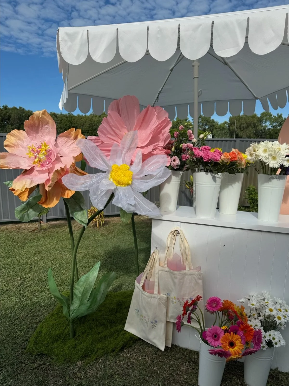 Flower stand under a white canopy with various pink, white, and orange flowers in white buckets and tote bags.