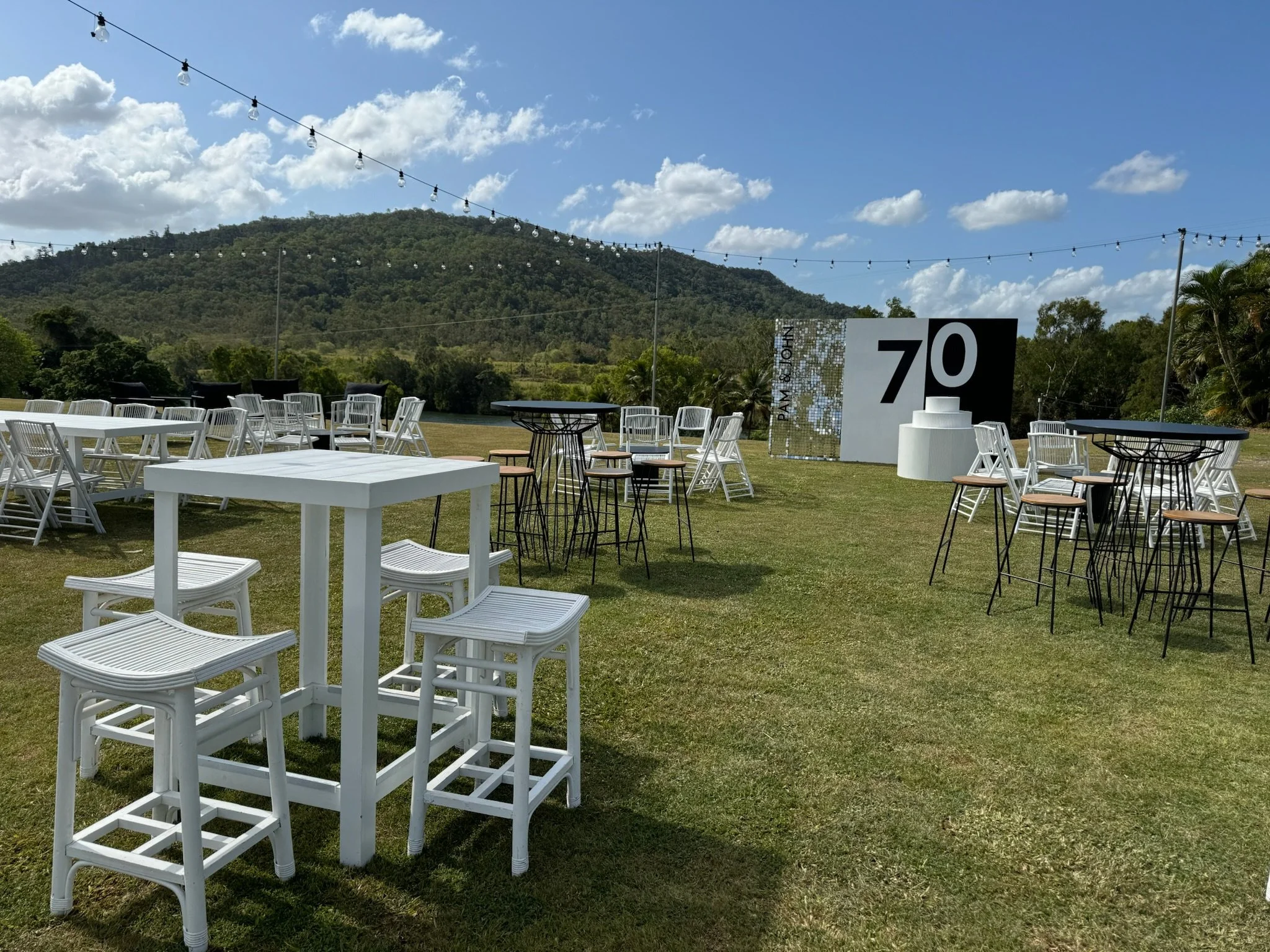 Outdoor event setup with white and black tables and chairs on a grassy field, with a backdrop of green hills, blue sky, and a large black and white sign displaying the number '70' and the name 'Pam & John'.