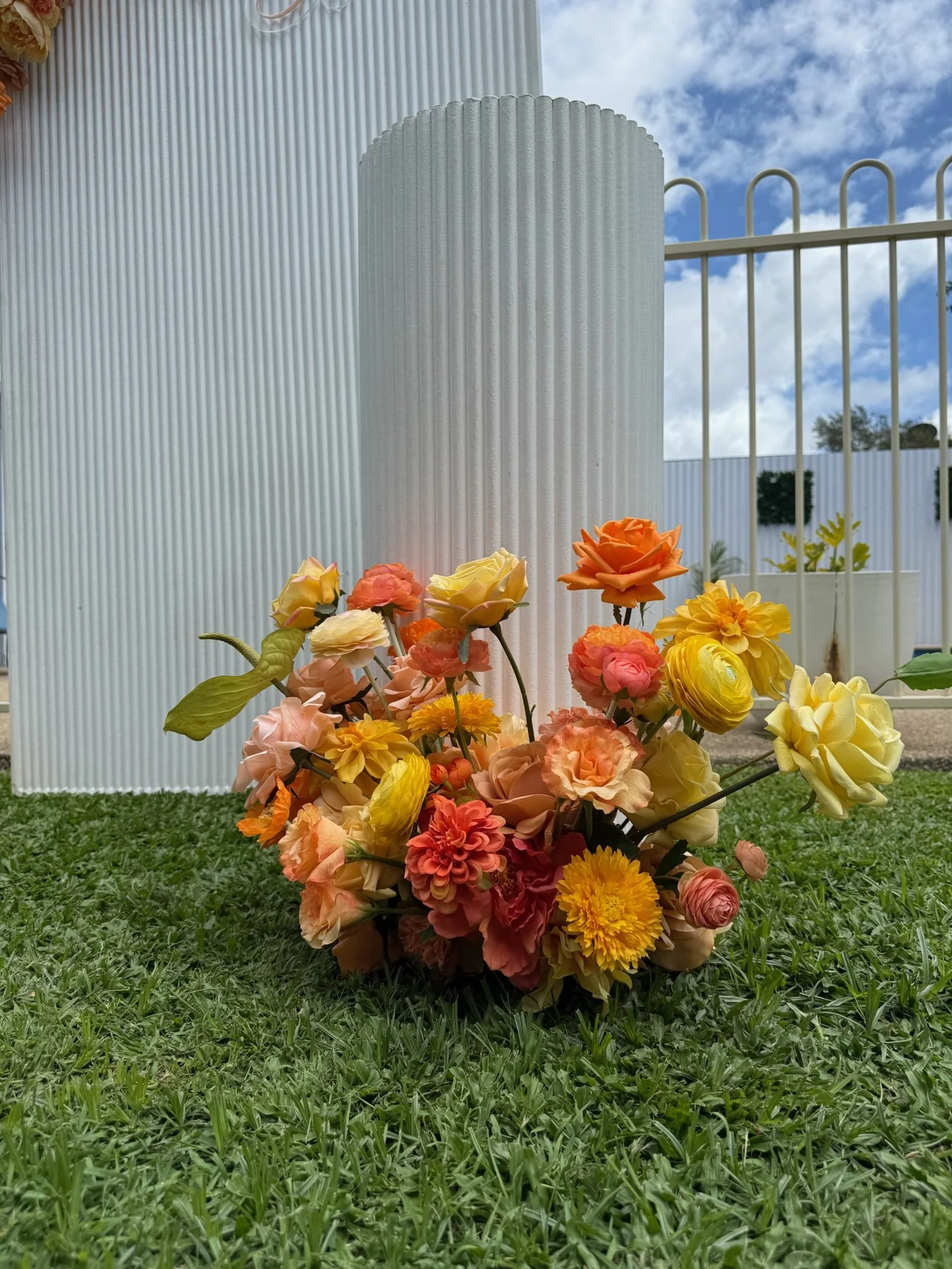 A colorful bouquet of artificial flowers, including roses and dahlias, resting on green grass outside near a white, textured cylindrical structure and a white fence, with a partly cloudy sky in the background.