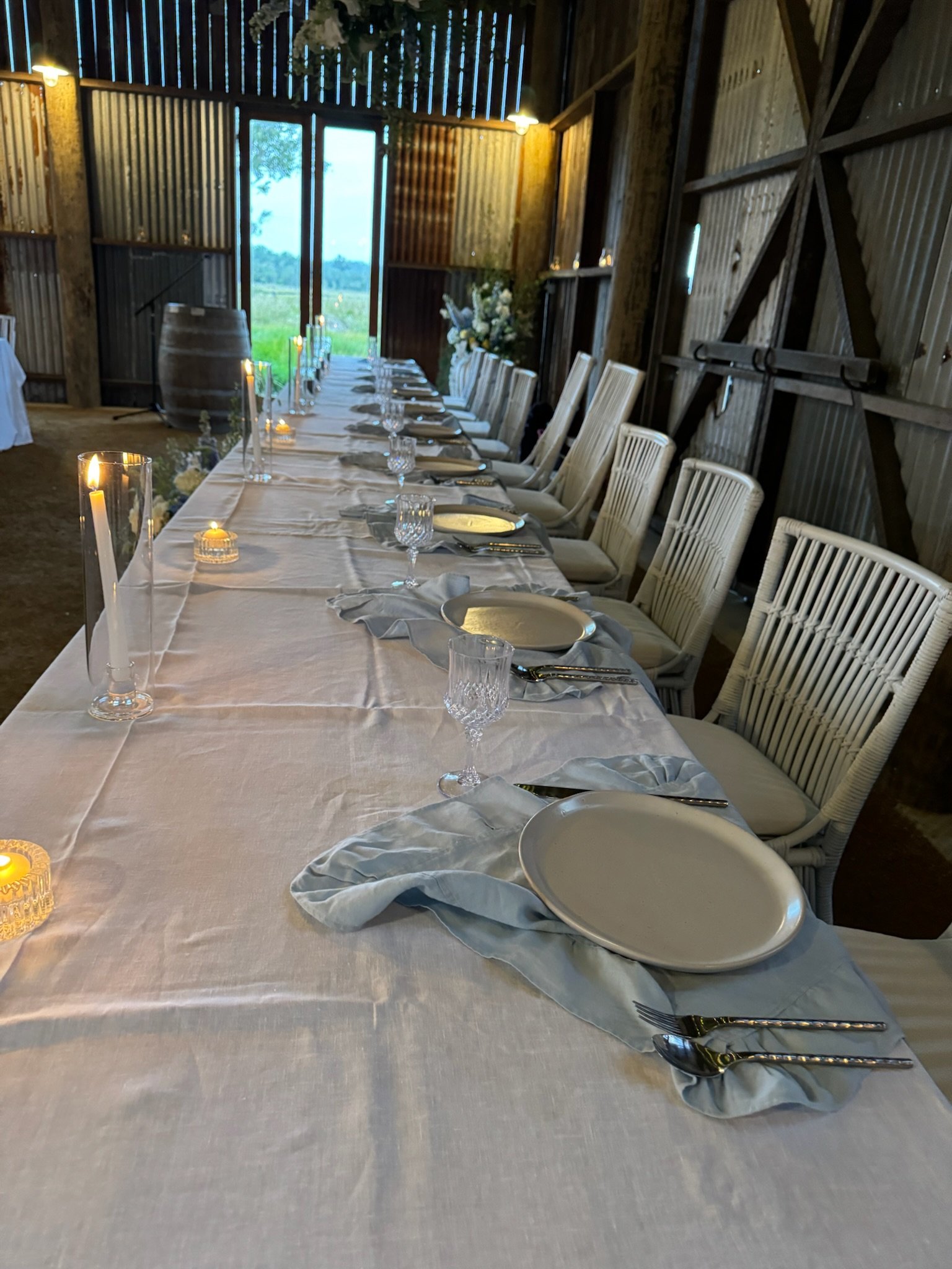 Long dining table set with white tablecloth, plates, silverware, glasses, candles, and floral arrangements in a rustic barn setting with open doors revealing greenery outside.