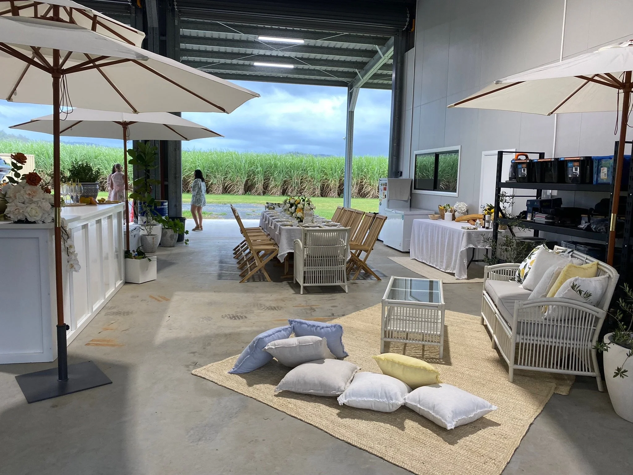Indoor space set up for an outdoor gathering with a long dining table, chairs, pillows, and umbrellas, with open doors revealing a grassy field and cloudy sky outside.