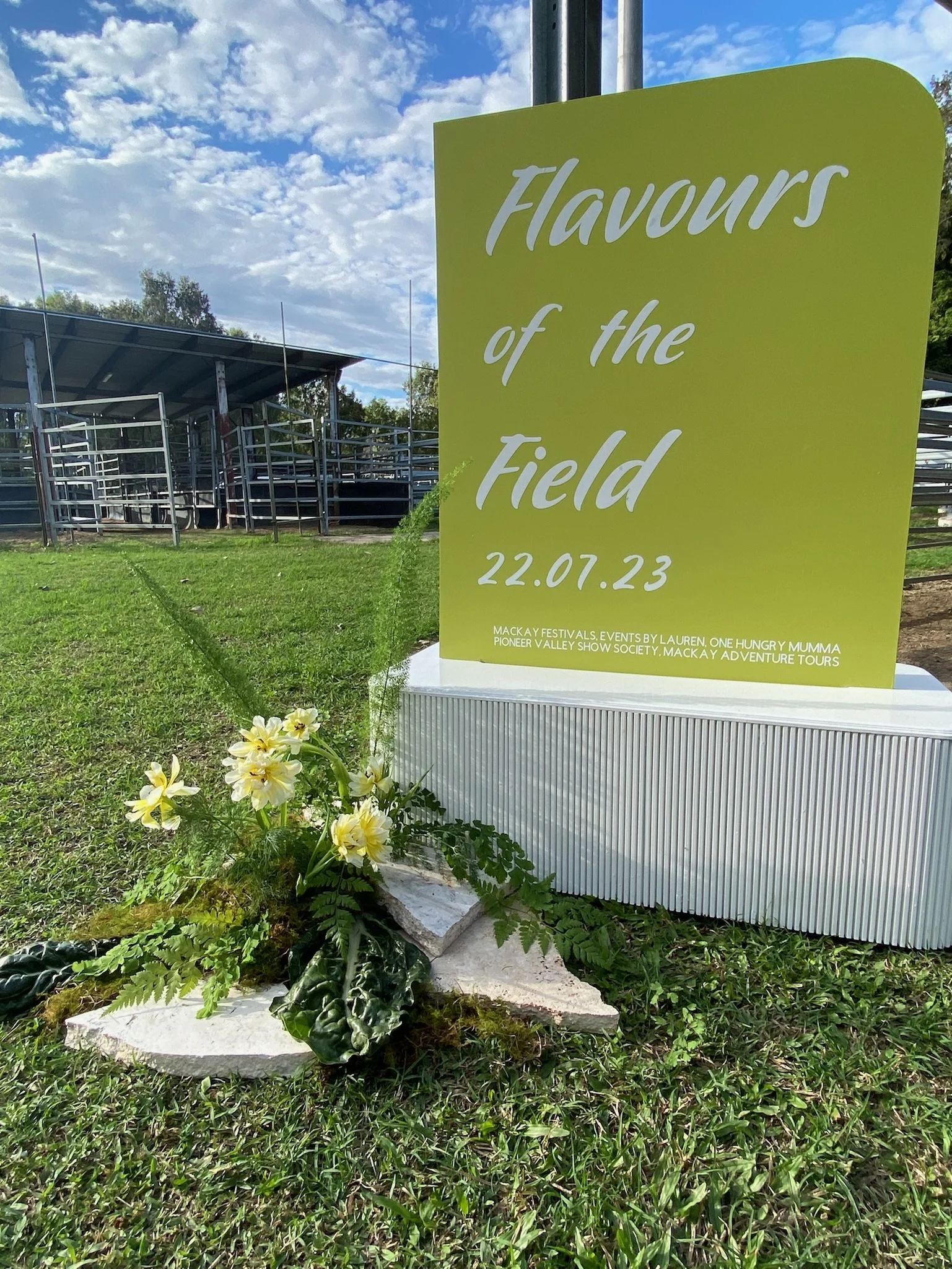 A bright green sign with white and light green lettering reads "Flavours of the Field" with the date 22.07.23, placed on a grassy field. Next to the sign, there is a small arrangement of yellow and white flowers with green foliage on white stones. In