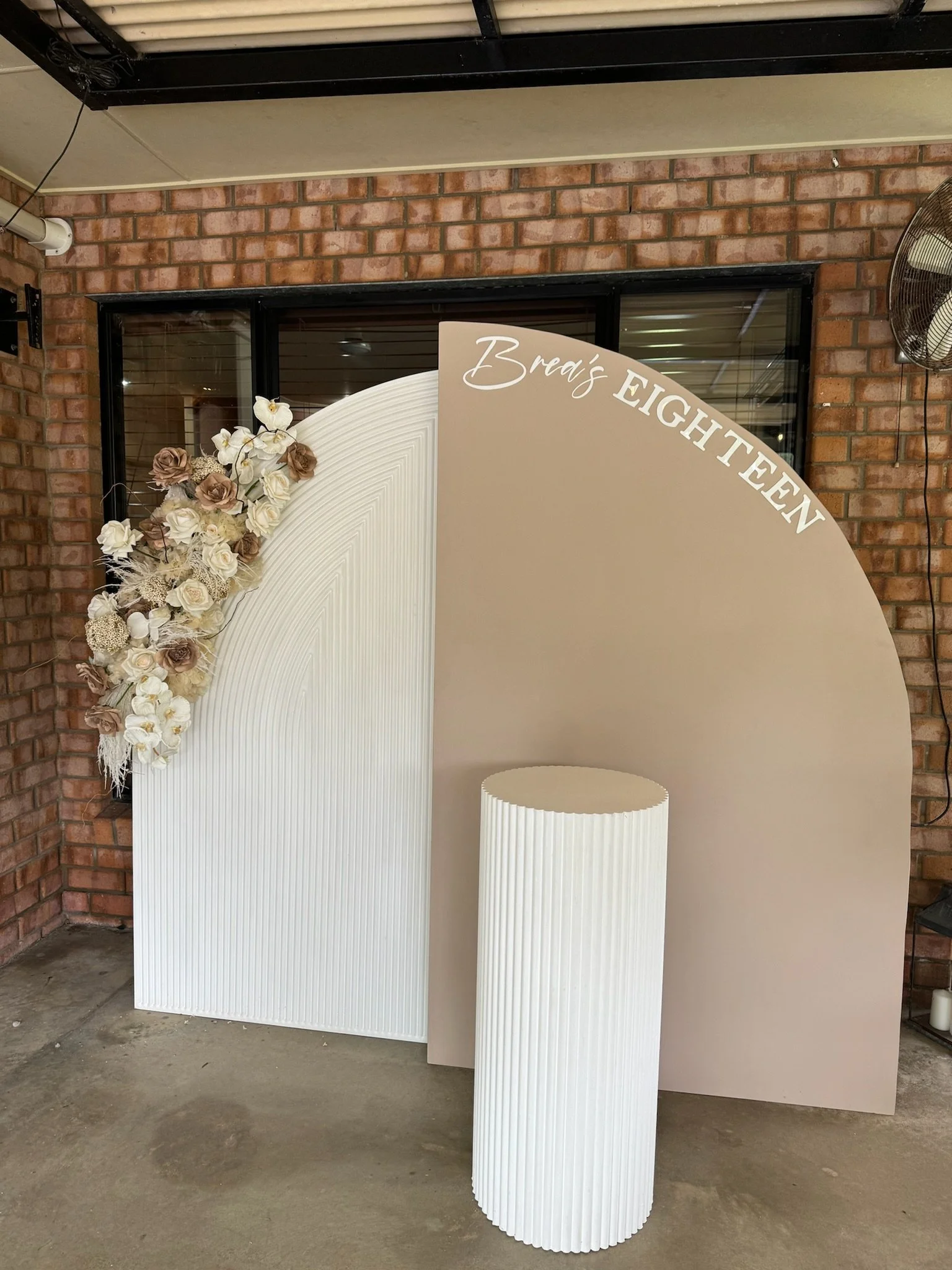 Celebration backdrop with a beige and white color scheme, decorated with white and beige flowers on the left side. The backdrop reads "Brea's EIGHTEEN" in white and beige lettering, indicating an 18th birthday celebration. There is a white, ribbed cy