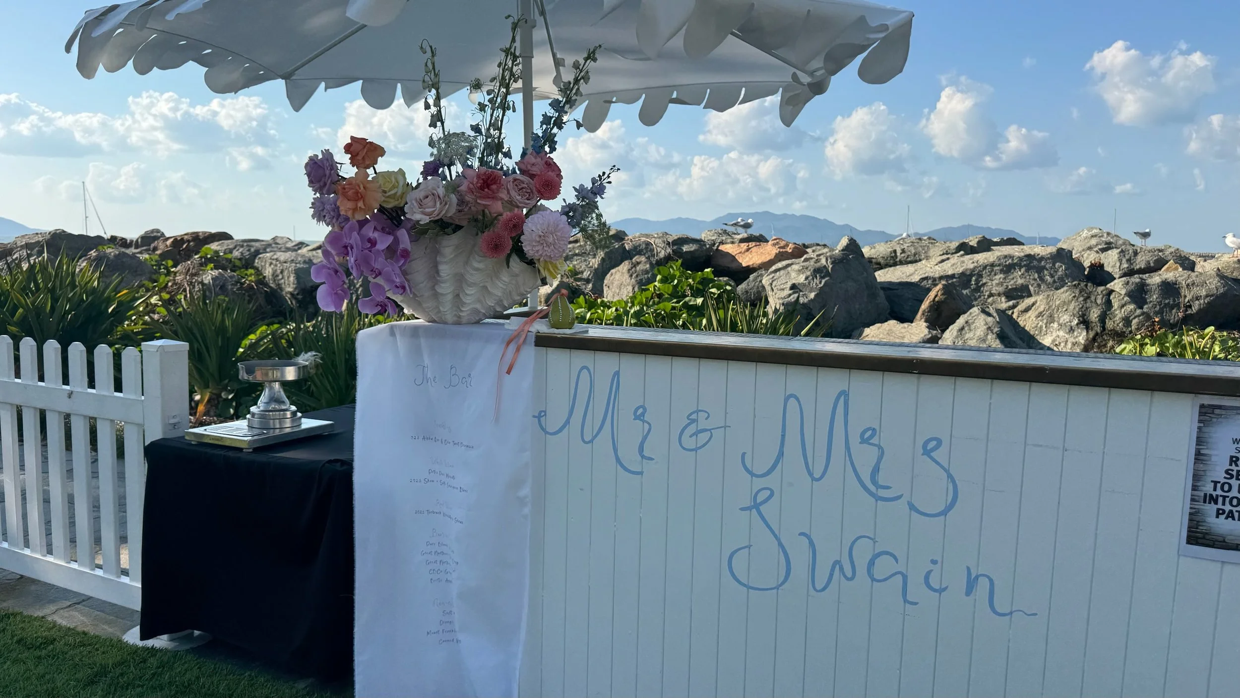 A seaside event setup with a white bar under a white umbrella, decorated with a large floral arrangement of pink, purple, and white flowers. The backdrop features rocks, the ocean, and a blue sky with clouds.