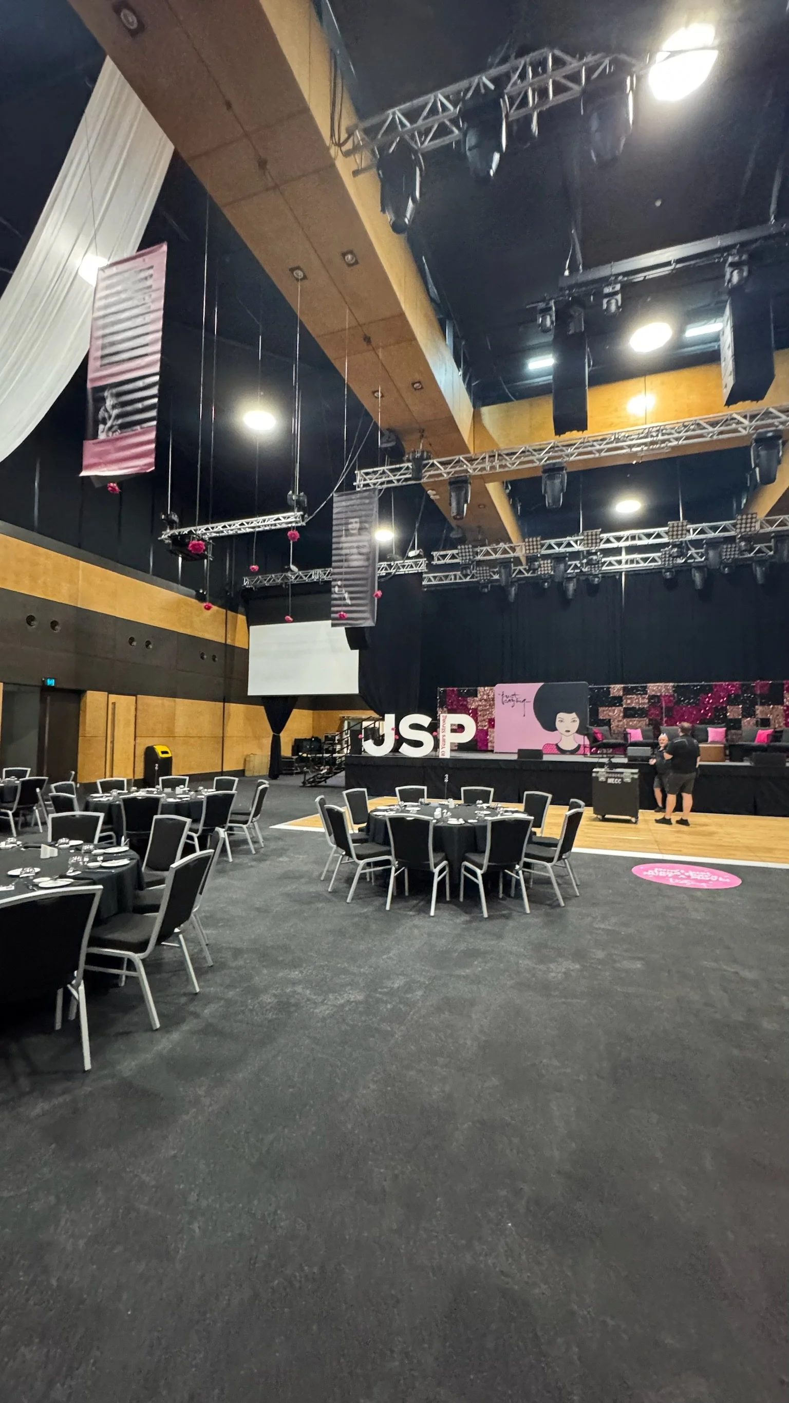 Event stage decorated with pink and black floral background and decorations, set up with large white letters "USP"; tables with black tablecloths and chairs in front of the stage, indicating preparation for a formal event or conference.
