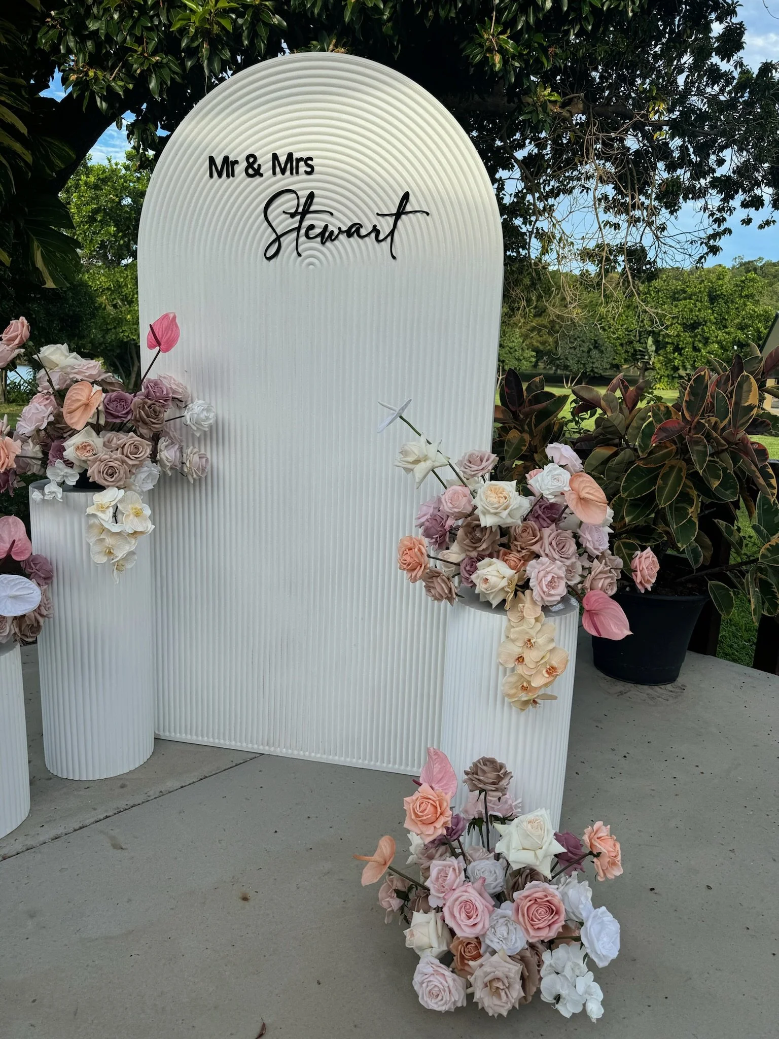 A wedding or event display with a white, ribbed backdrop reading 'Mr & Mrs Stewart' in black and cursive, surrounded by pastel-colored flower arrangements on white vases and a black pot, set outdoors with trees and a blue sky in the background.