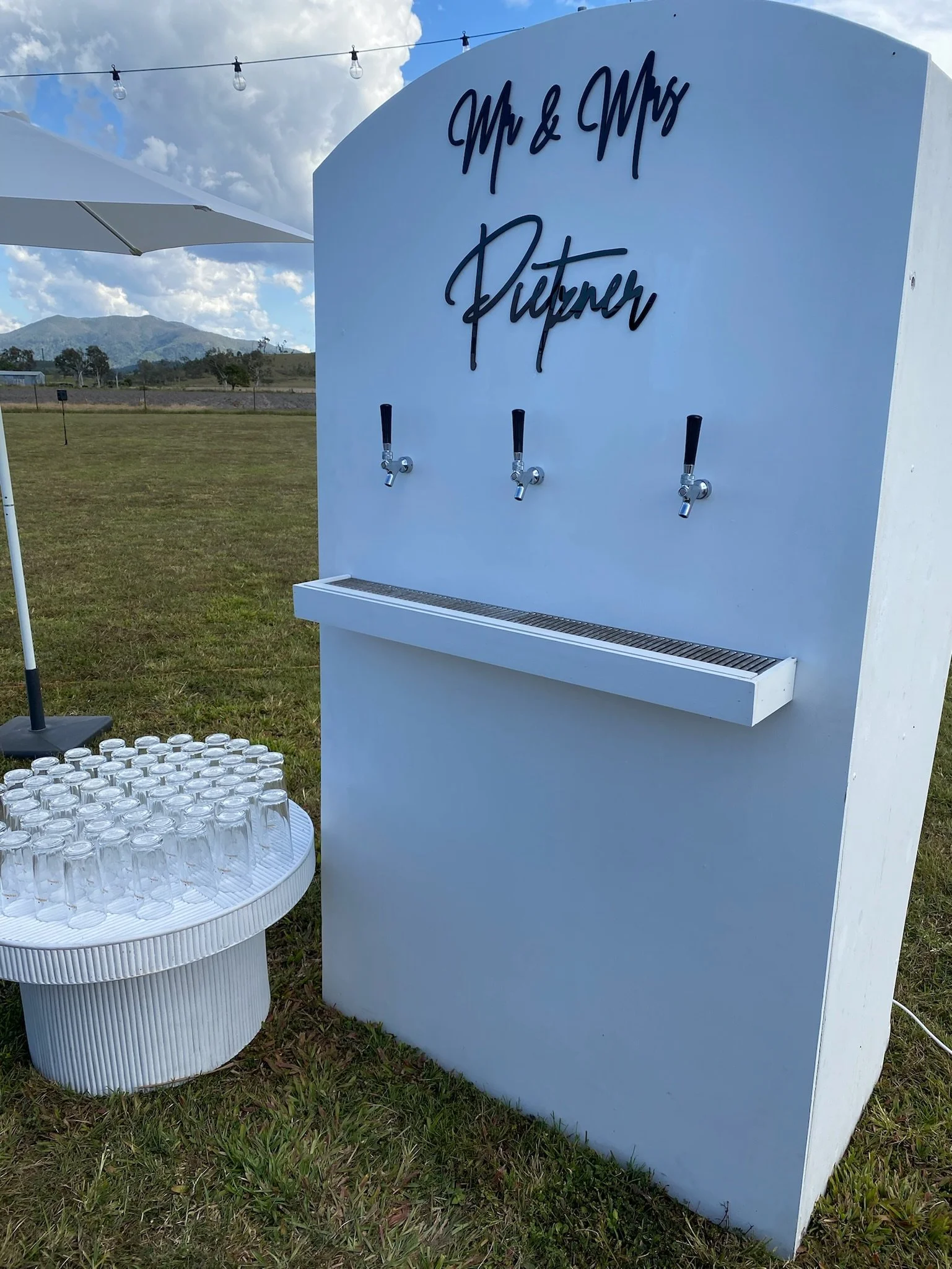 White outdoor drink dispenser with three taps, labeled 'Mr & Mrs Putney' at a wedding or outdoor event. To the left, a round table with multiple upside-down glasses. A white umbrella and a landscape with mountains are in the background.