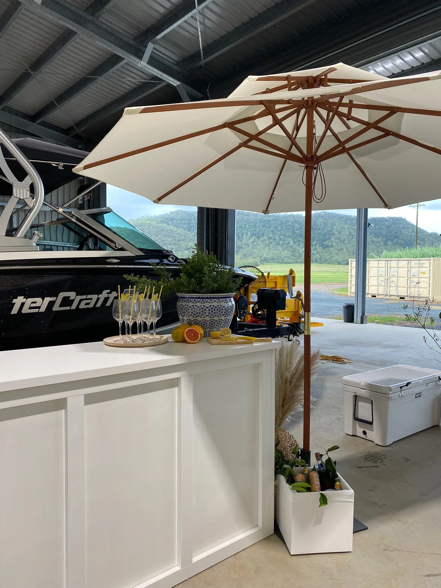 A white bar counter with a potted plant, grapefruit, and lemons, topped with glasses of drinks with yellow straws, under a large white patio umbrella in a garage with a boat and outdoor scenery in the background.