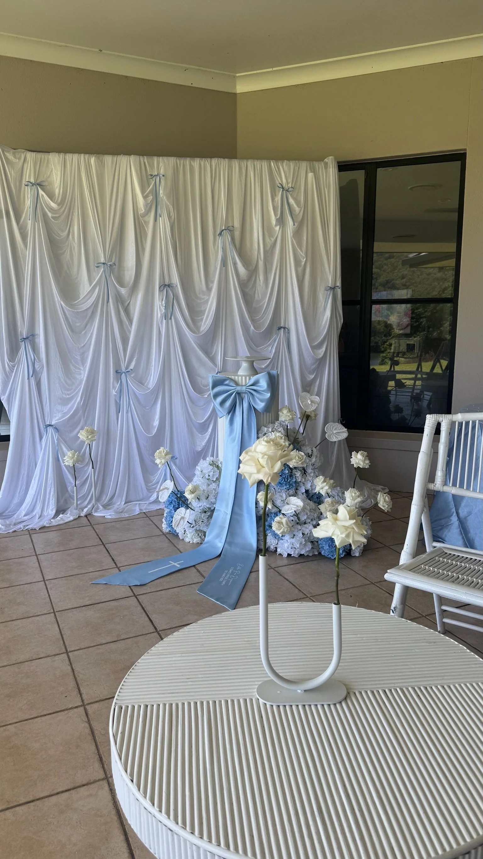 Decorative setup for a wedding with a white draped backdrop, blue bows, and white flowers on the floor.