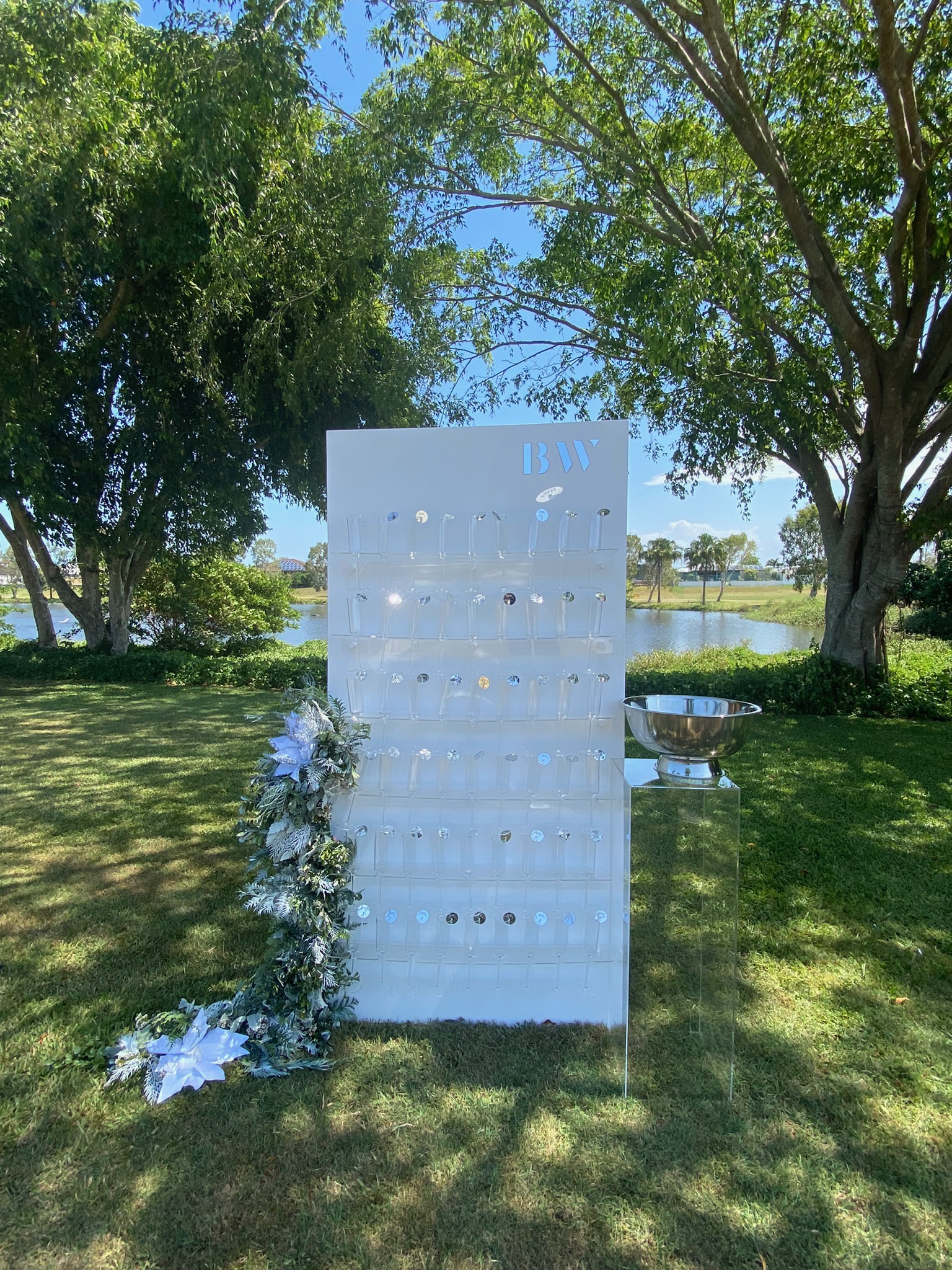 Outdoor wedding or event decor featuring a white stand with hooks, a floral arrangement with white poinsettias, a silver bowl on a clear stand, trees, a grassy lawn, a pond, and a blue sky.