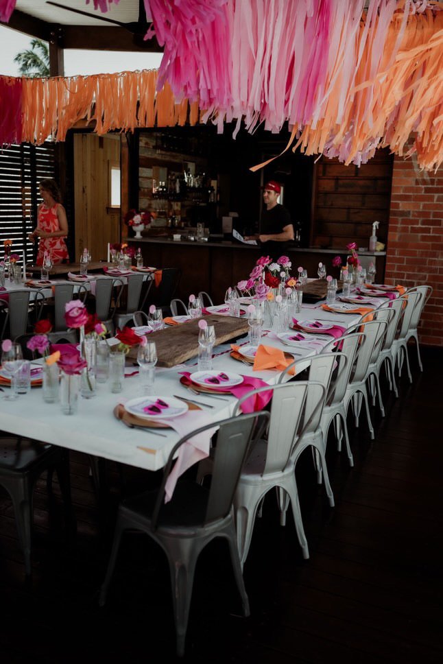Decorated dining table with pink and orange accents, set for a celebration, under colorful hanging paper decorations, with a bar and a person working behind it.