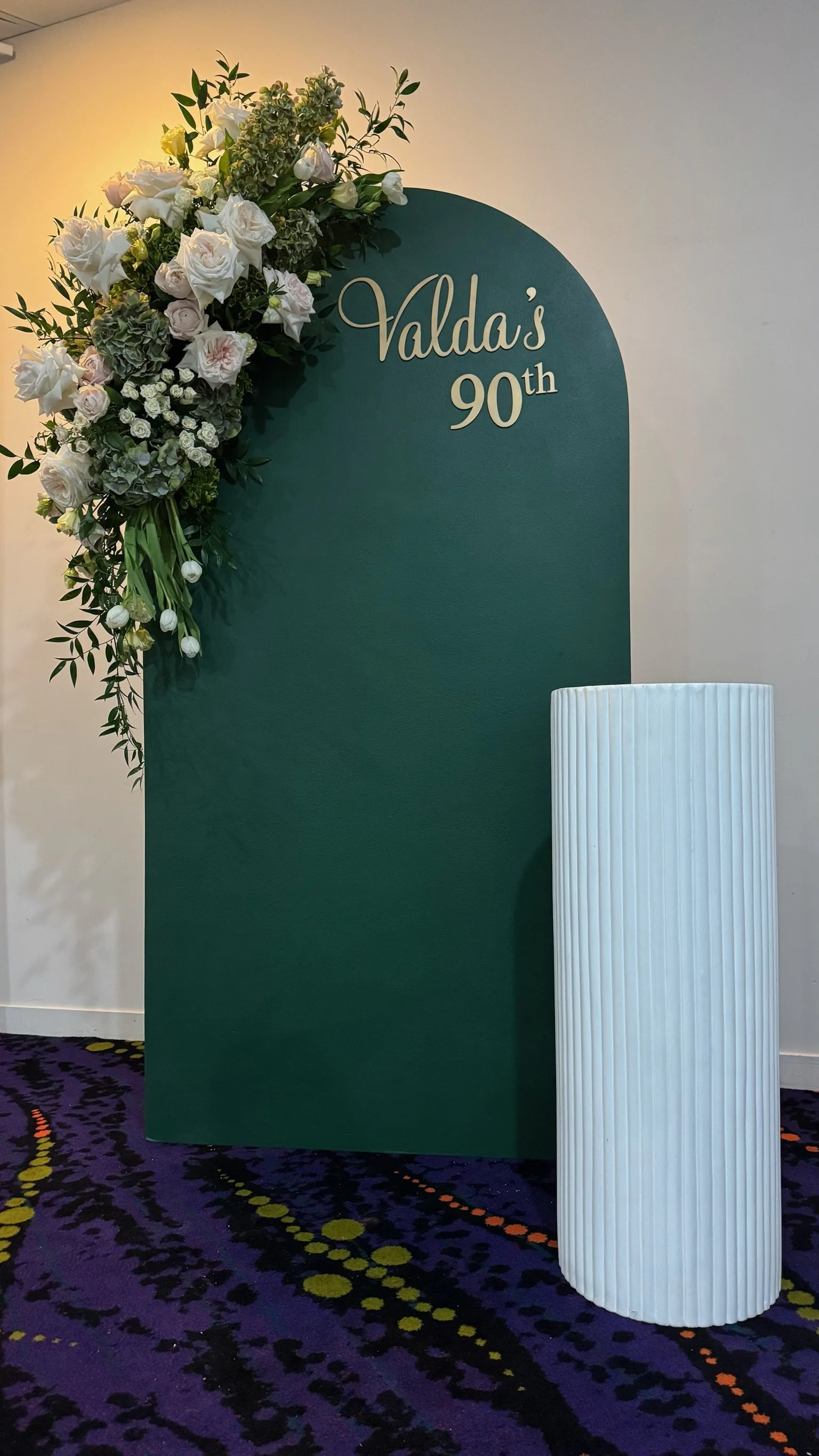 A decorative display with a dark green arched panel, decorated with white and green floral arrangements on the top left, and text that reads "Valda's 90th" in white cursive and bold print. A white cylindrical vase with vertical ribbing stands to the 