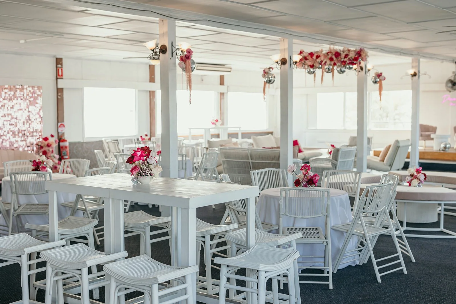 Decorated event space with white tables, chairs, and pink flower centerpieces, adorned with pink and white floral arrangements and hanging decorations.