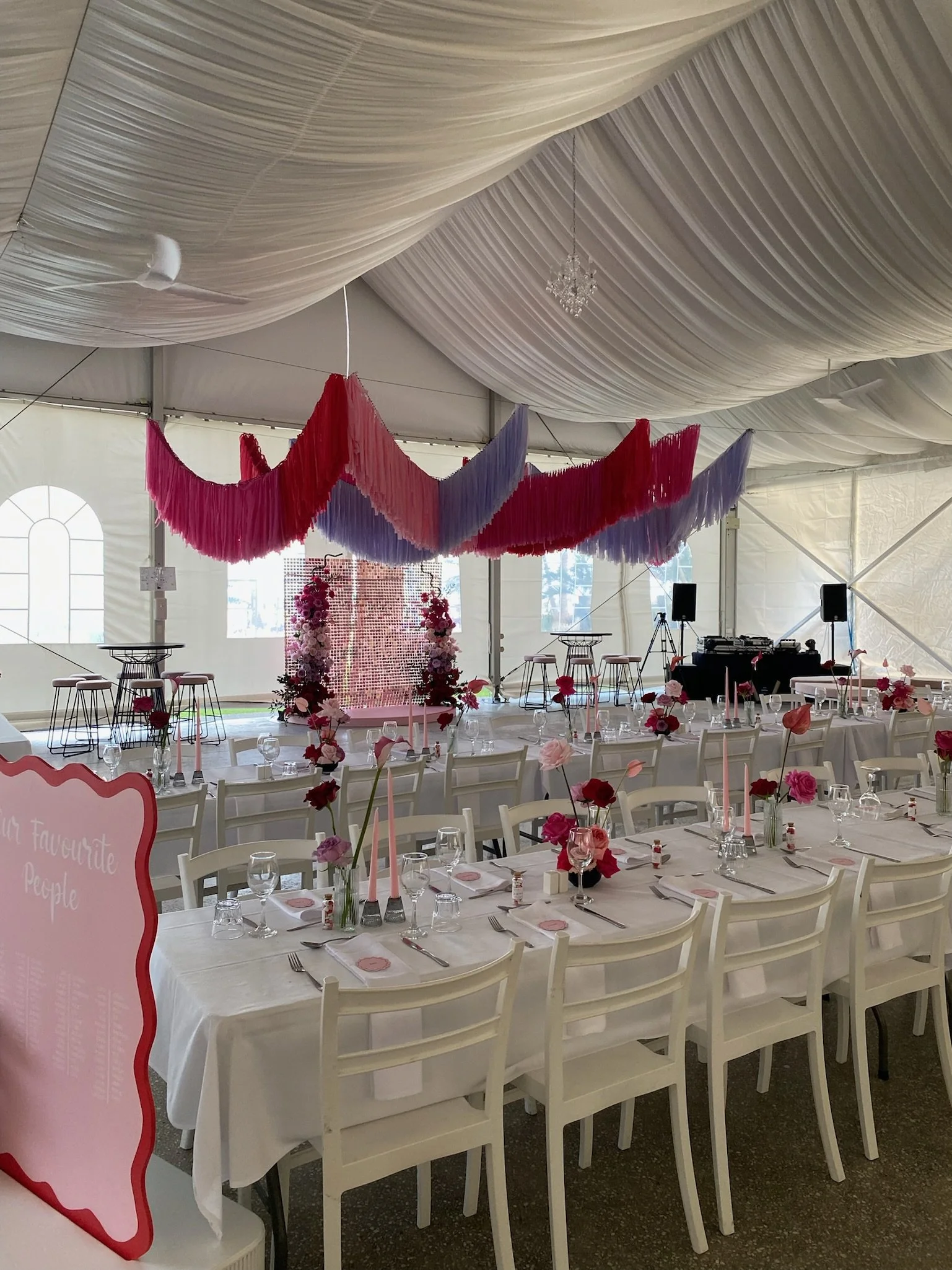 Inside a decorated event tent with a long dining table set with white tablecloths, pink and red flowers, pink candles, and glassware. The ceiling has draped fabric with pink, purple, and red hanging decorations, and there's a small stage at the back 