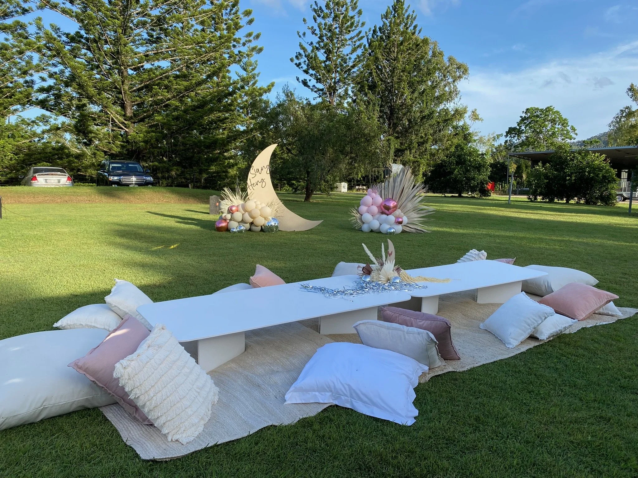 Outdoor celebration setup with low white table surrounded by pillows on a grassy lawn, with decorative installations of balloons and plants in the background.