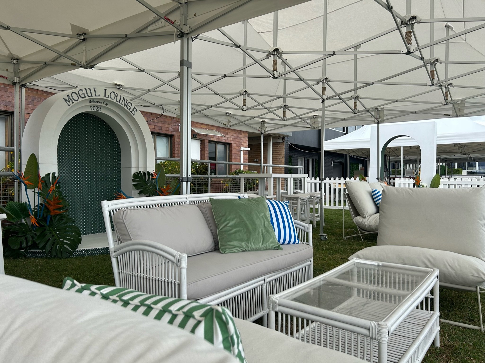Outdoor lounge area with white wicker furniture, cushions, and pillows under a large white tent, near a brick building with a sign reading 'Mogul Lounge' and plants with orange flowers.