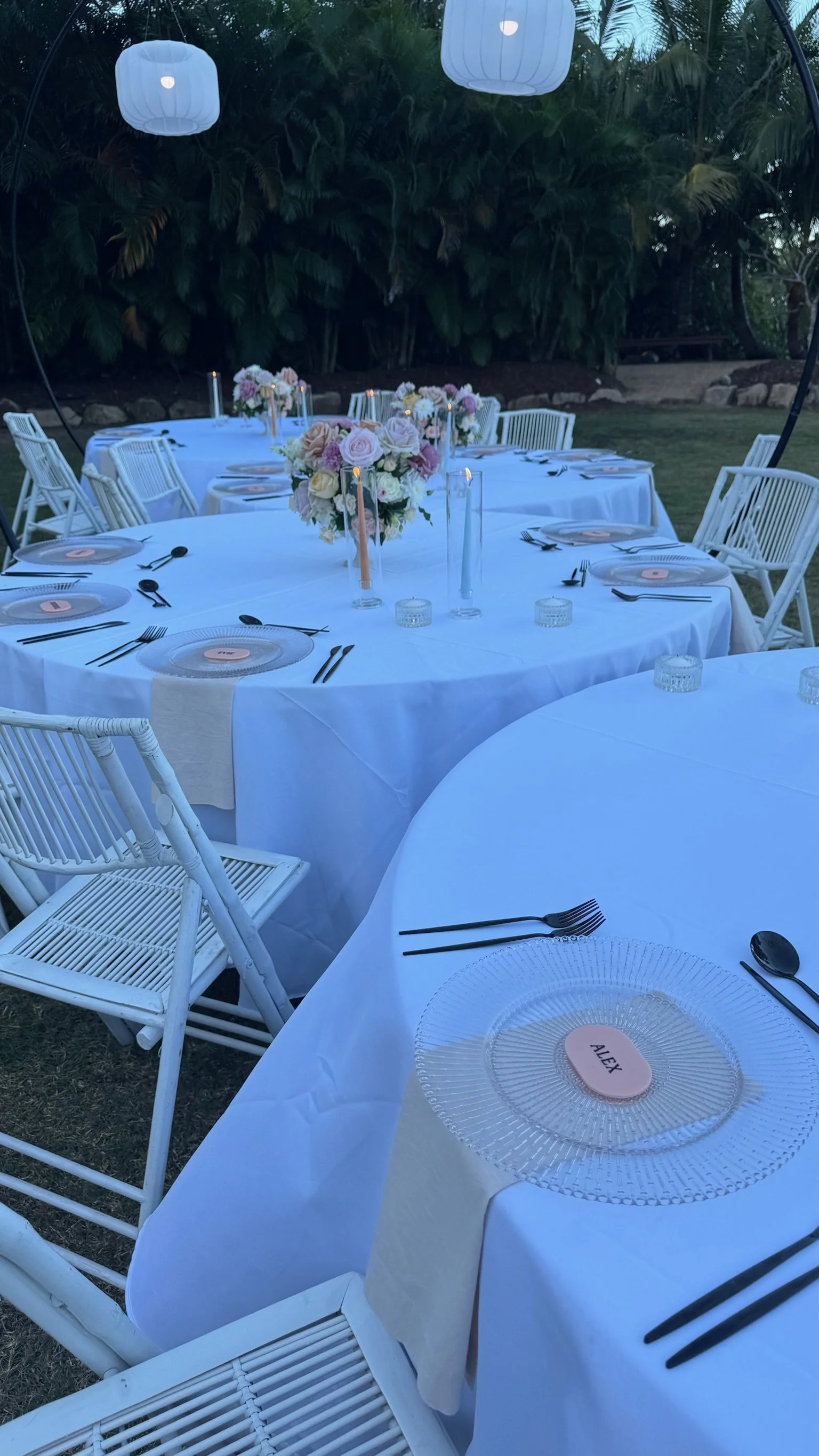 Outdoor event setup with round tables covered in white tablecloths, decorated with pink and white floral centerpieces and tall candles. White chairs surround the tables, and the scene is lit by hanging lanterns with a backdrop of green foliage.