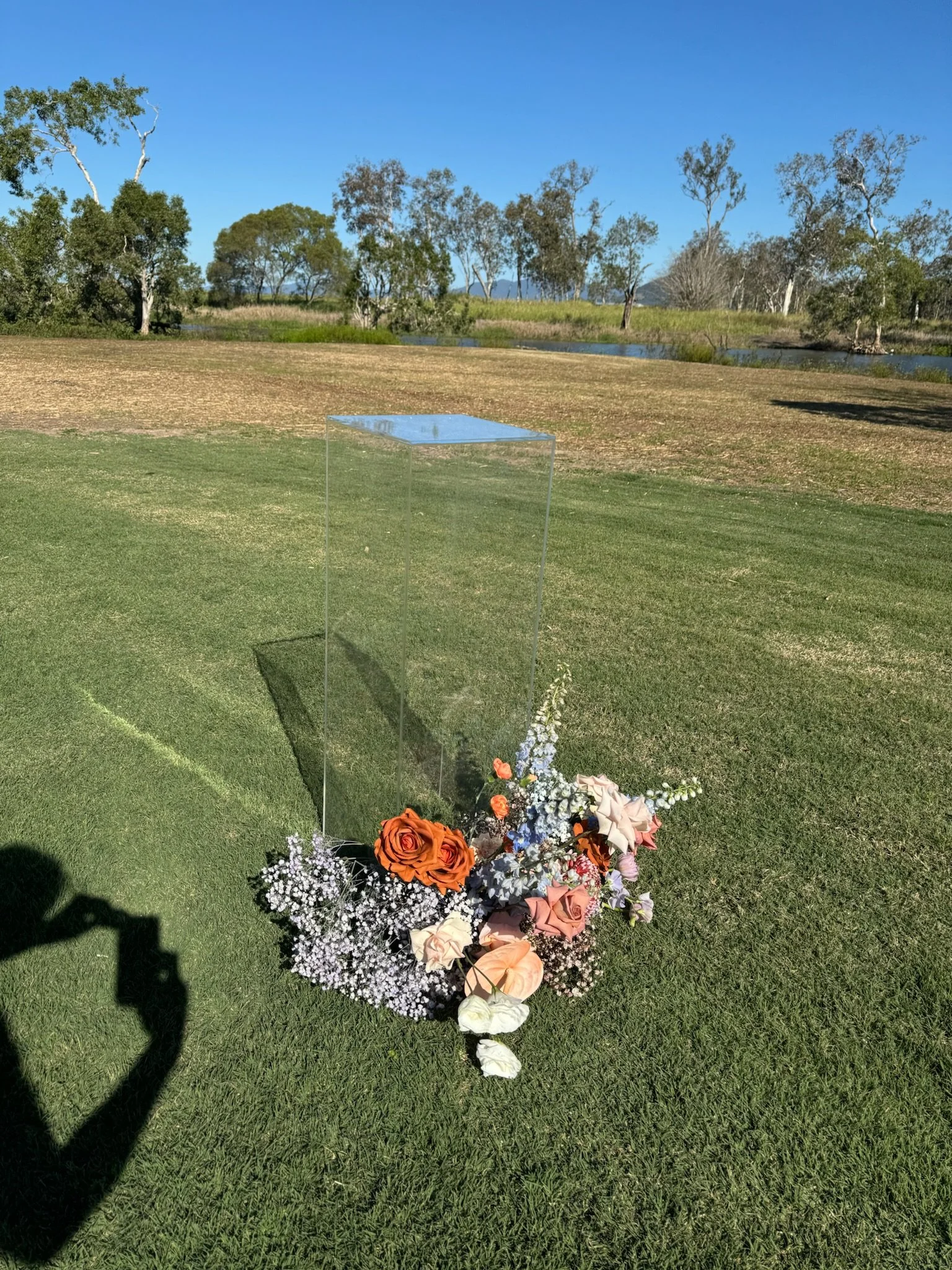 A clear acrylic memorial or display case placed on green grass with a border of mixed pink, orange, white, and blue flowers at the base. In the background, there is a landscape with trees, a body of water, and a bright blue sky.