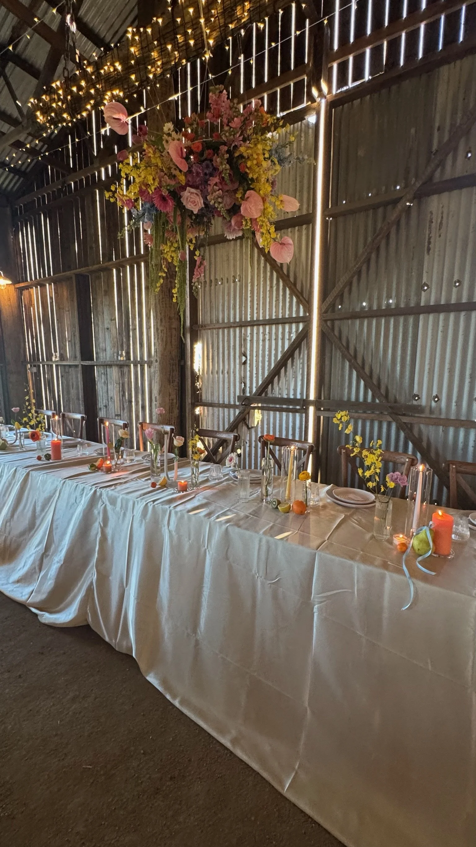 Long banquet table with white tablecloth, decorated with small vases of colorful flowers, candles, and scattered fruits, set against a rustic wooden barn interior with string and fairy lights, and a large floral arrangement hanging on the wall.