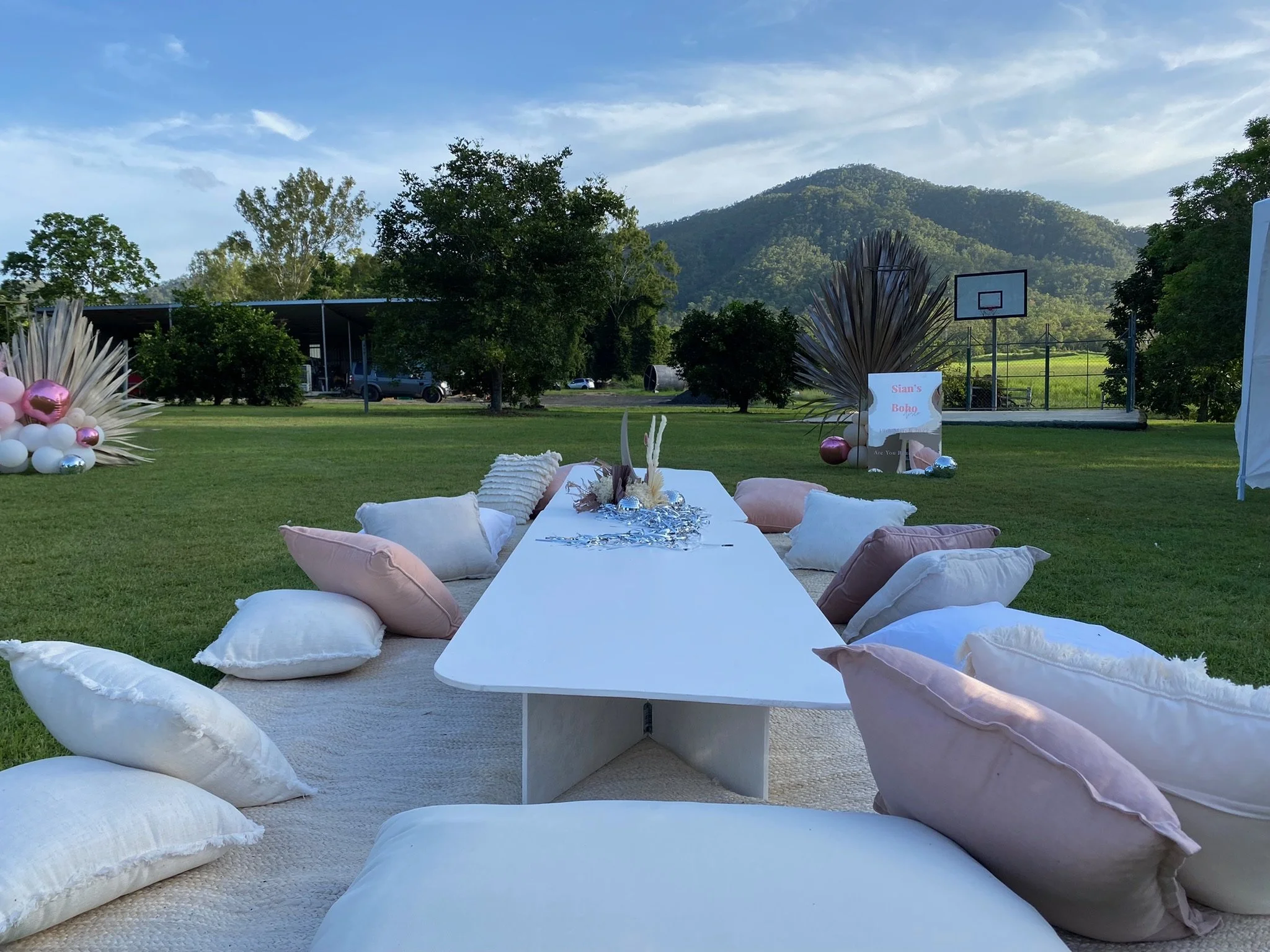 Outdoor setup with a long white table surrounded by pillows, with a backdrop of trees, a mountain, and a fence, featuring decorative items and balloons.