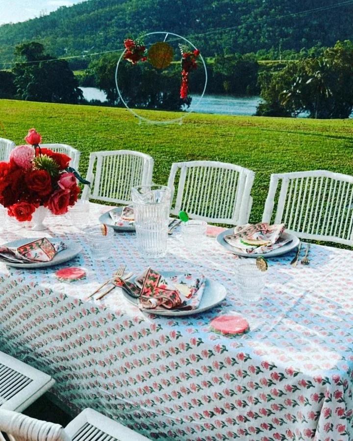 Outdoor dining table set with floral tablecloth, pink and red floral arrangement, pink napkins, clear glasses, and cutlery, with white chairs, green grass, trees, river, and hills in the background.
