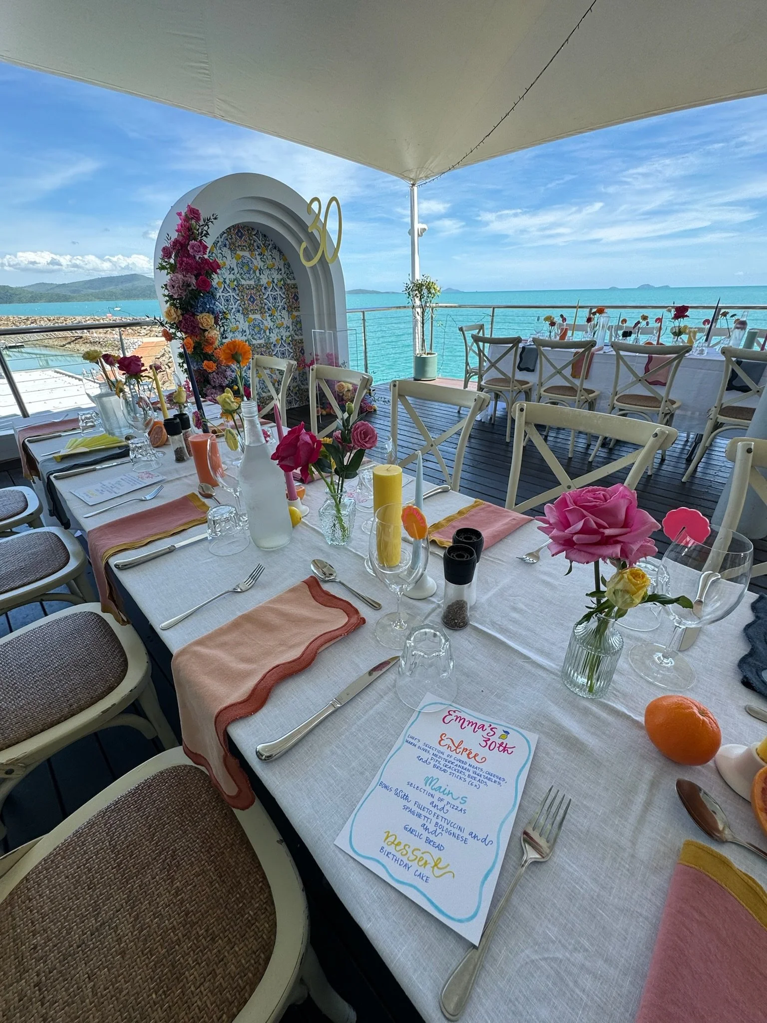 Overwater birthday celebration setup with ocean view, decorated tables, colorful flowers, candles, and a menu card.