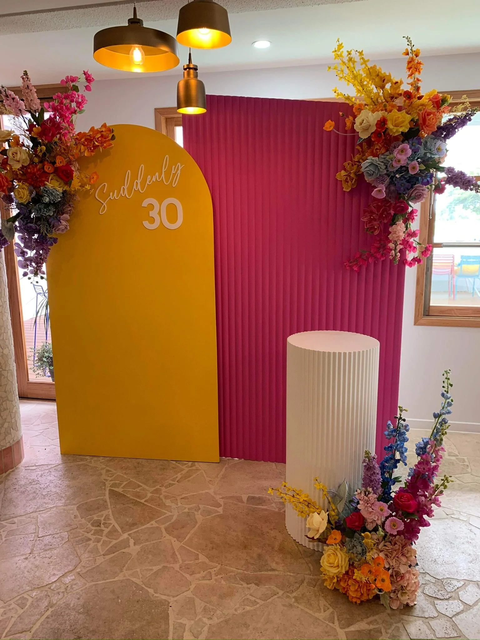 Colorful photo booth backdrop with a yellow arch stating "Suddenly 30" and decorated with vibrant flowers, pink folding screen, and a white fluted cylindrical pedestal.