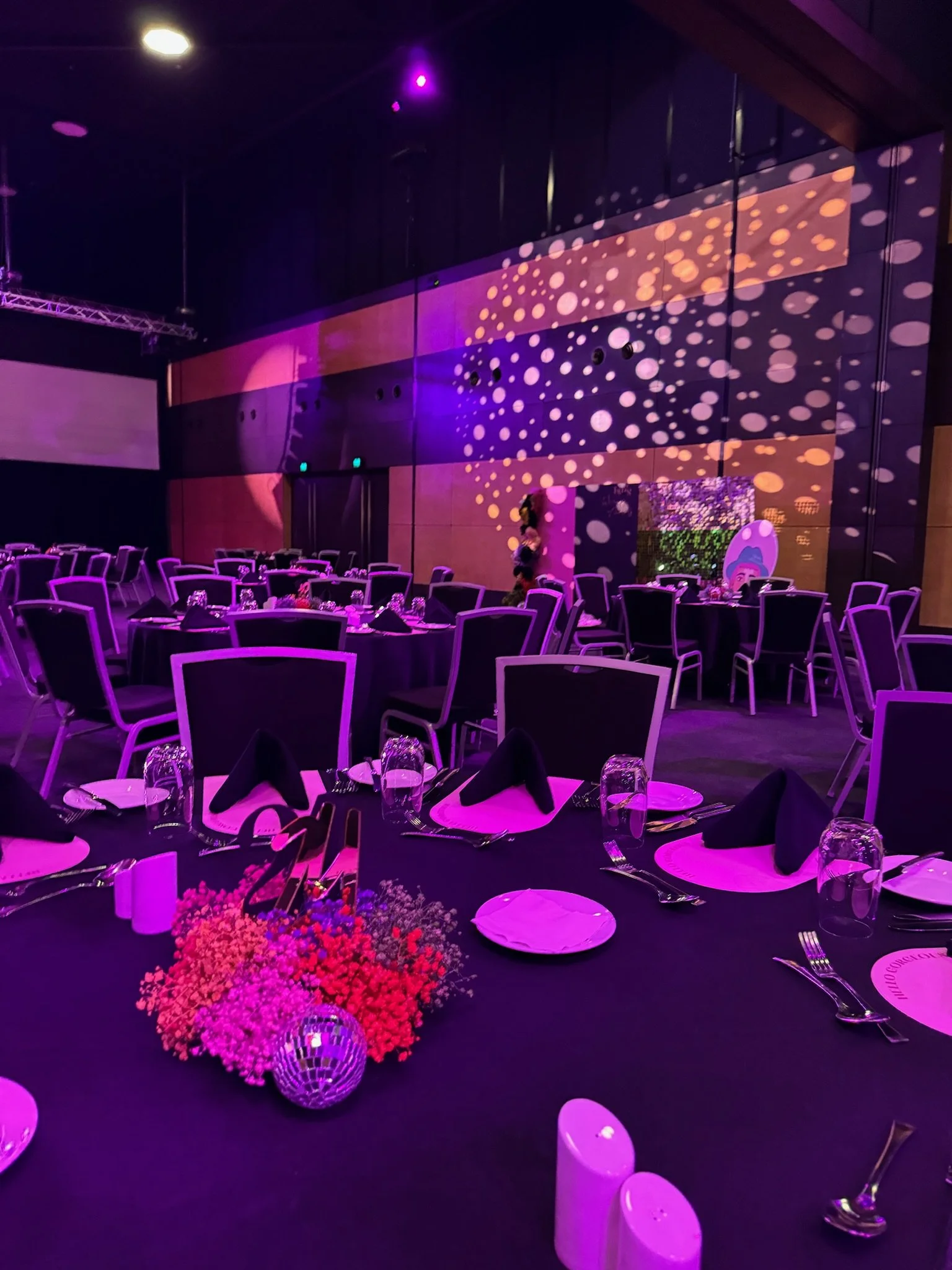 A banquet hall decorated for an event with multiple round tables covered in black tablecloths, black napkins, and glassware, accented with pink and purple lighting, and a floral centerpiece on each table.