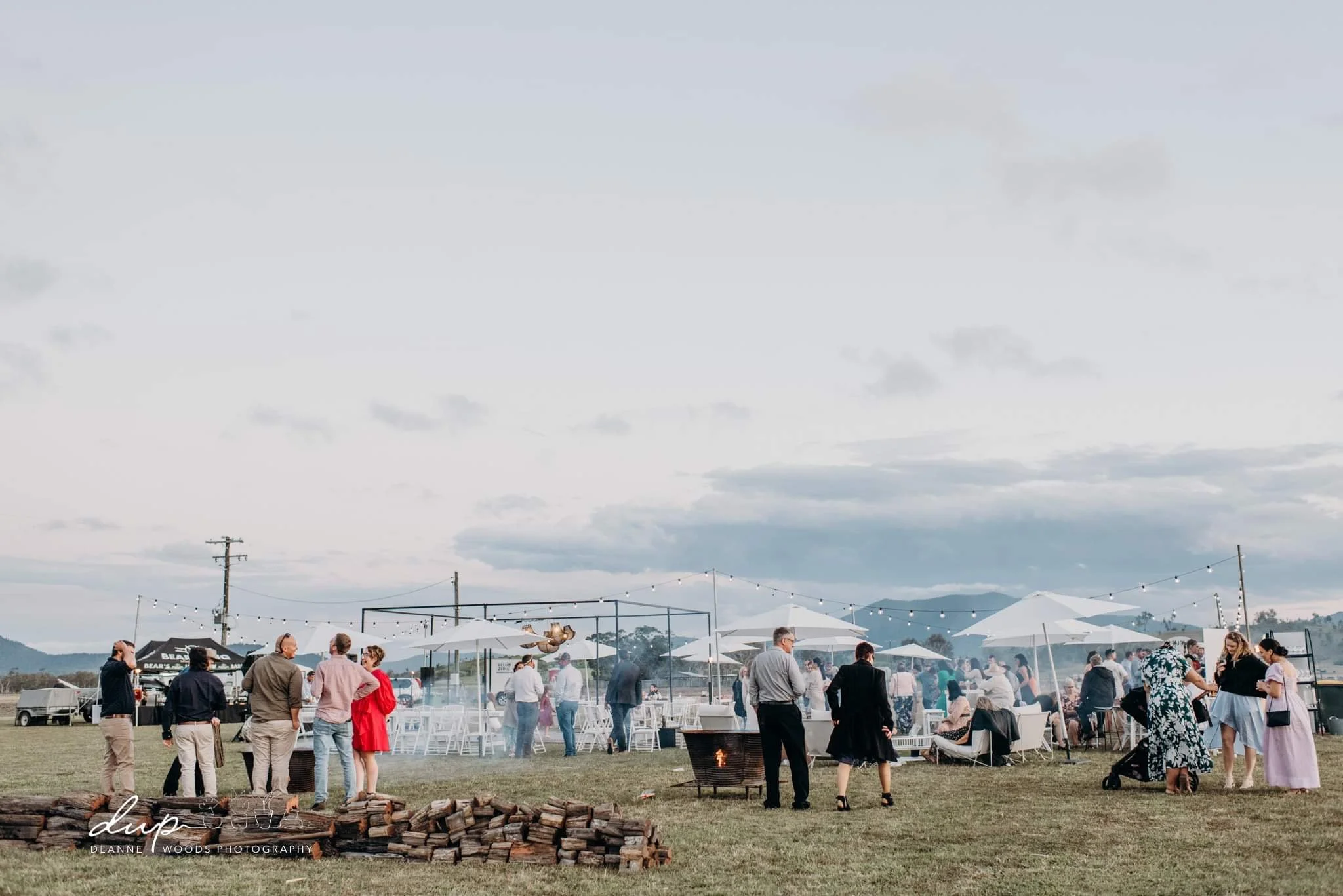 People gathered outdoors at a social event or celebration under large umbrellas with string lights, some standing near a fire pit, while others sit on chairs or stand in groups, with a grassy field and mountains in the distance.
